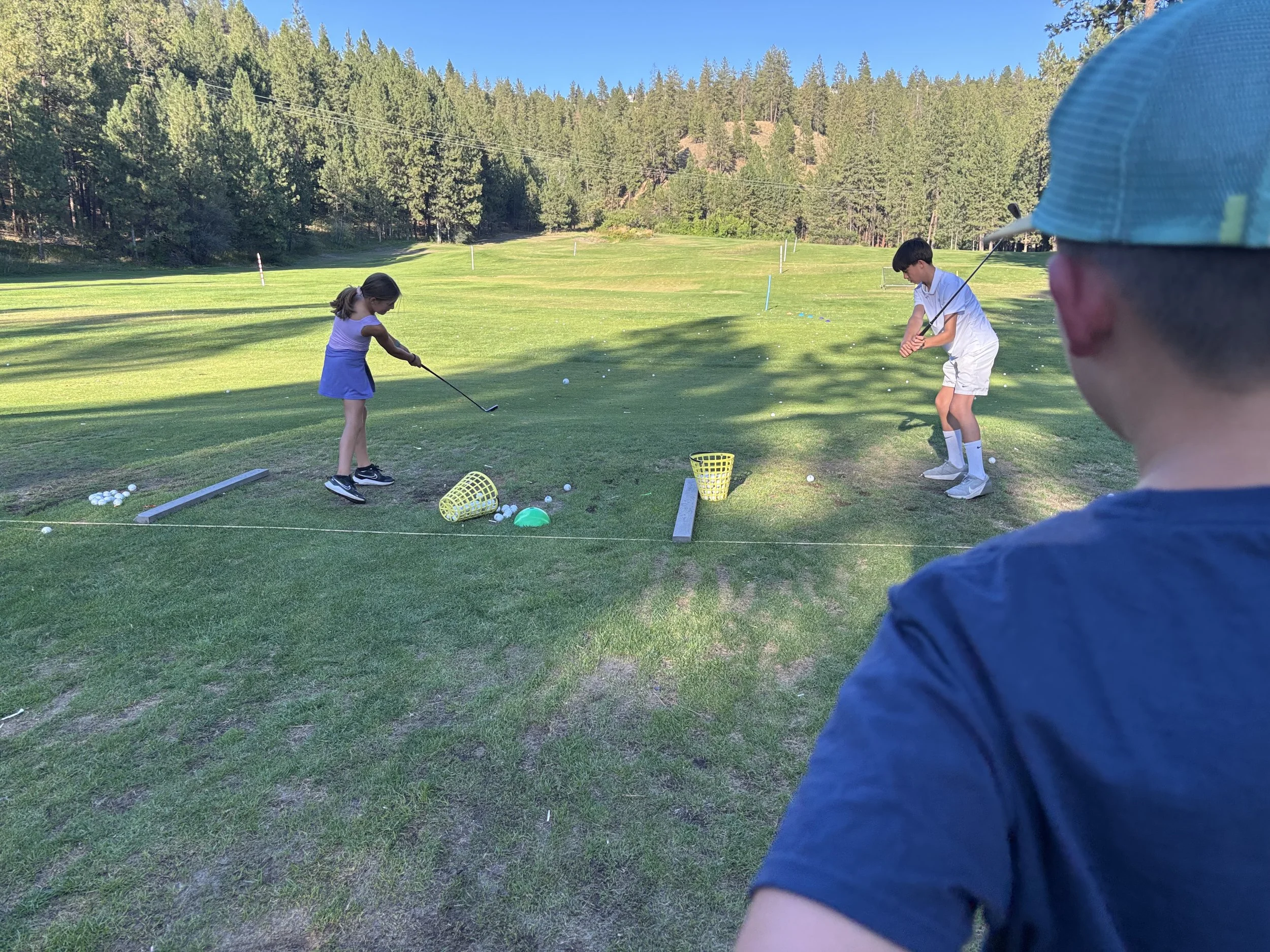 Two children playing golf on a practice range, with a person observing in the foreground, surrounded by golf balls and baskets, on a sunny day with trees and grass in the background.