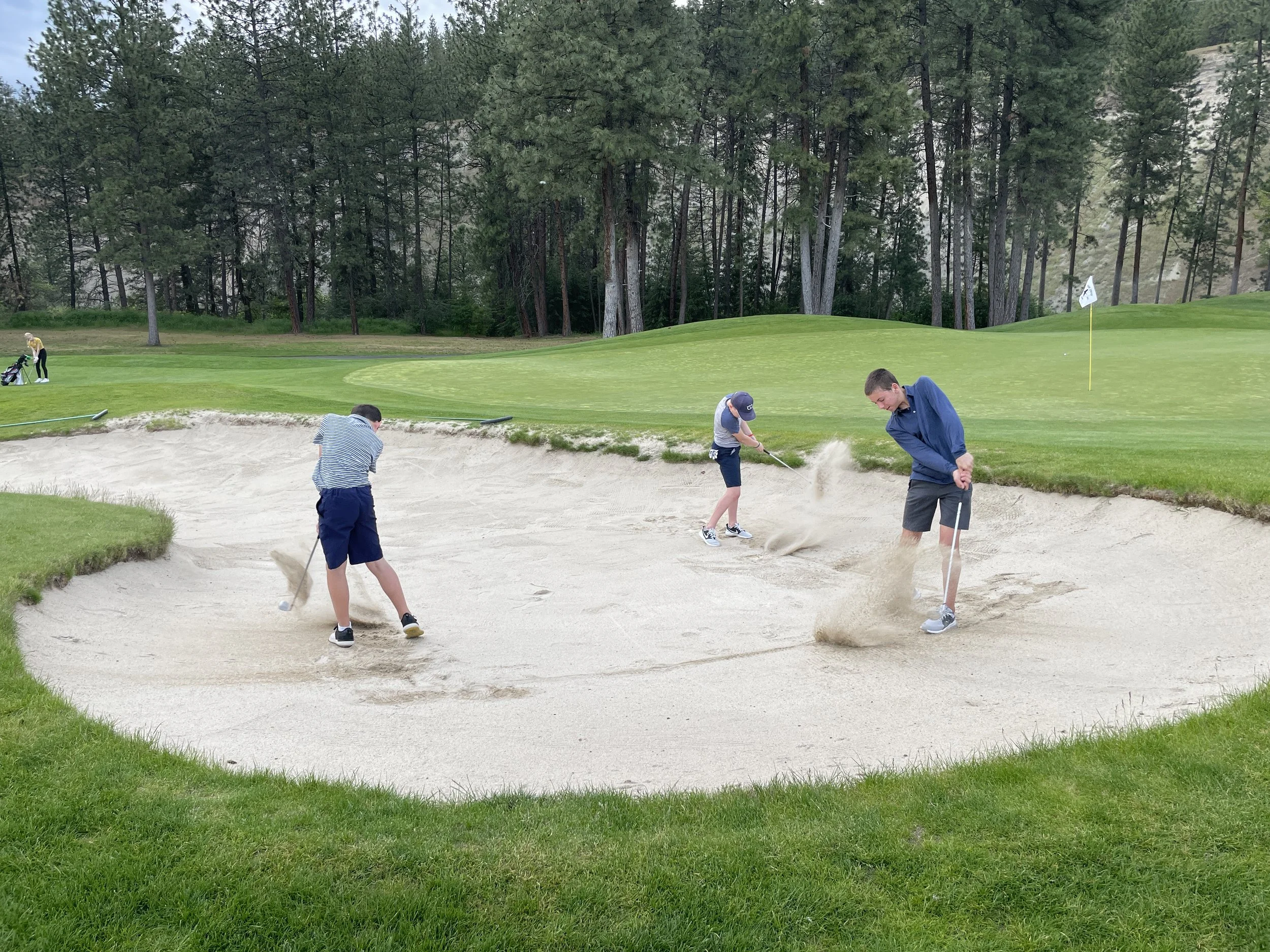 Three boys hitting golf balls from a sand bunker on a golf course, with a green and trees in the background.