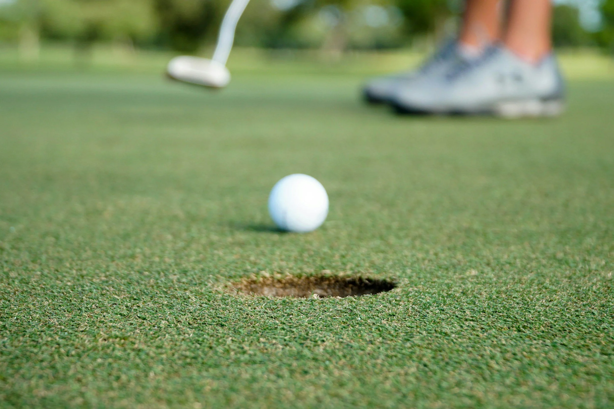 Close-up of a golf ball near a hole on a golf green, with a golfer's feet and golf club in the background.