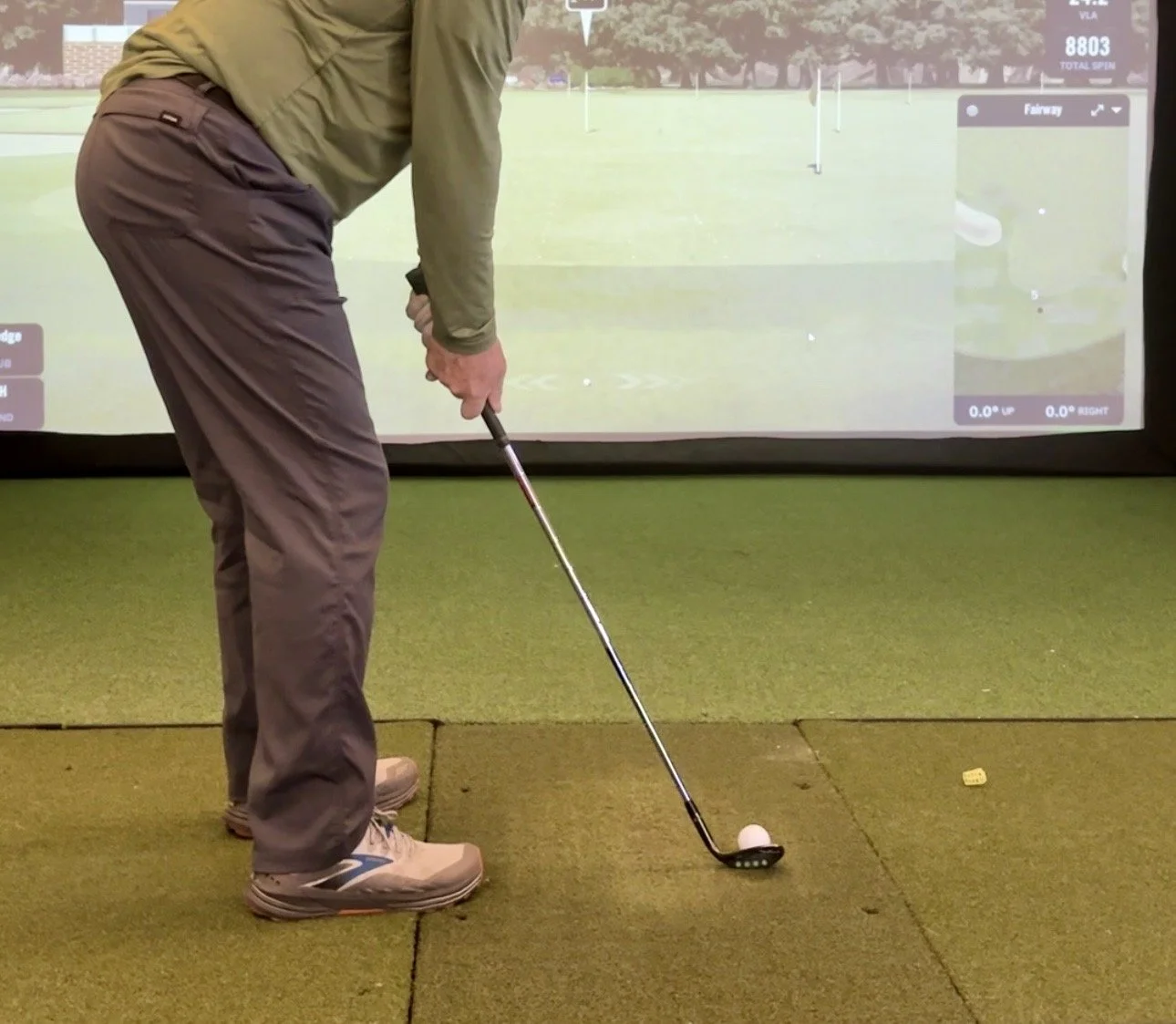Person practicing golf indoors, preparing to hit a golf ball on a synthetic mat with a golf simulator in the background