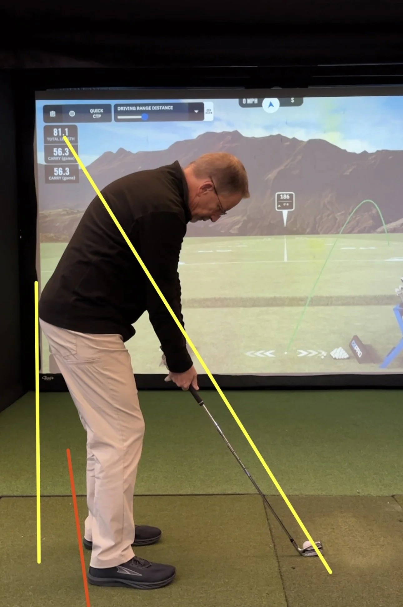 Man in black jacket and beige pants practicing golf swing in indoor simulator with golf screen in background.