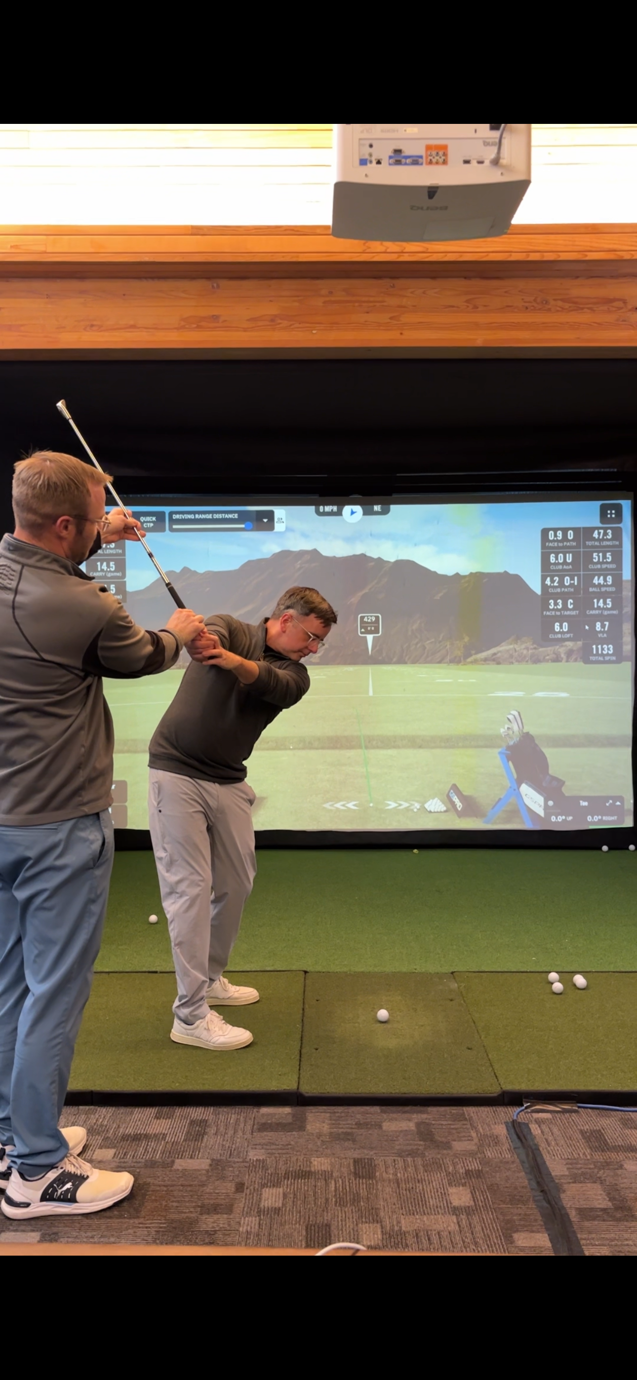 Two men practicing golf swings in a golf simulation room with a large screen displaying a virtual golf course landscape.
