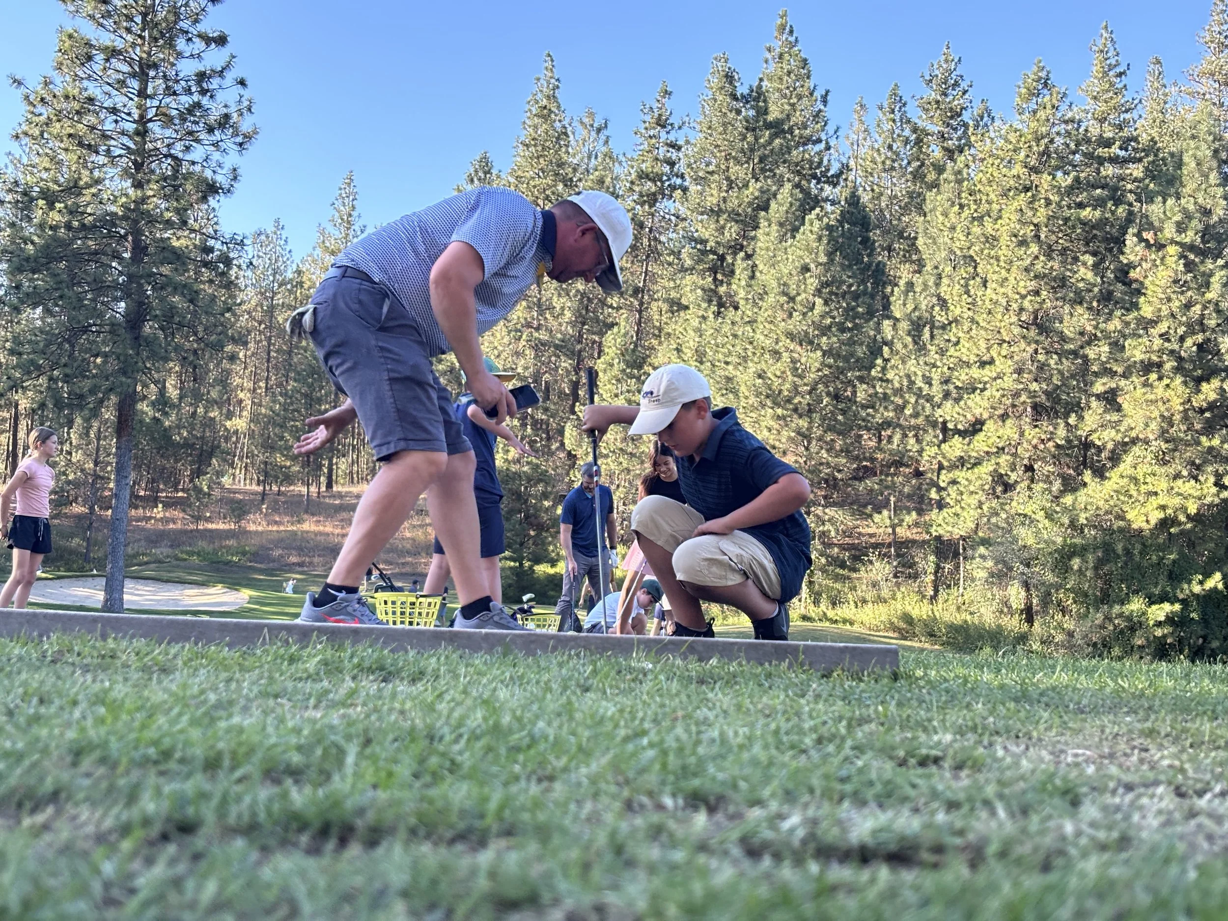 A man and a boy kneeling on the golf course, engaged in a game or activity, with several other people in the background near the green and trees.
