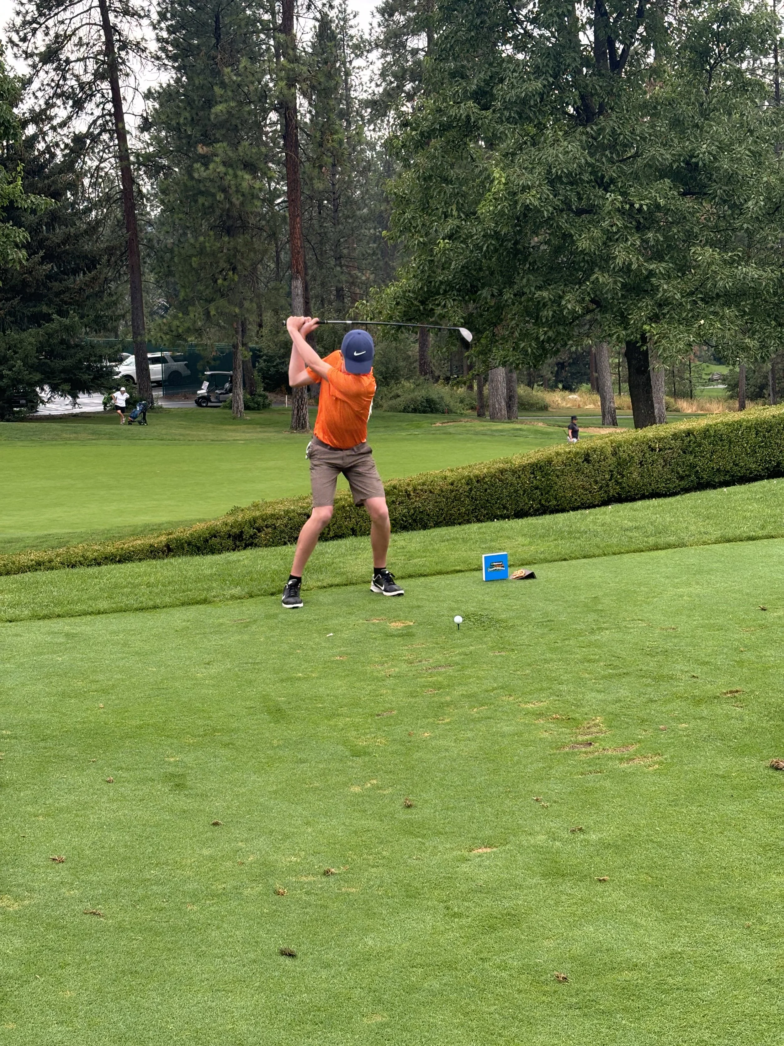 A person in an orange shirt and shorts swinging a golf club on a golf course.