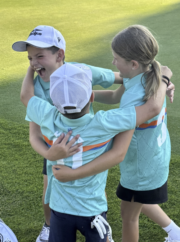 Three children in teal sports jerseys and white caps celebrating and hugging on a grassy field.