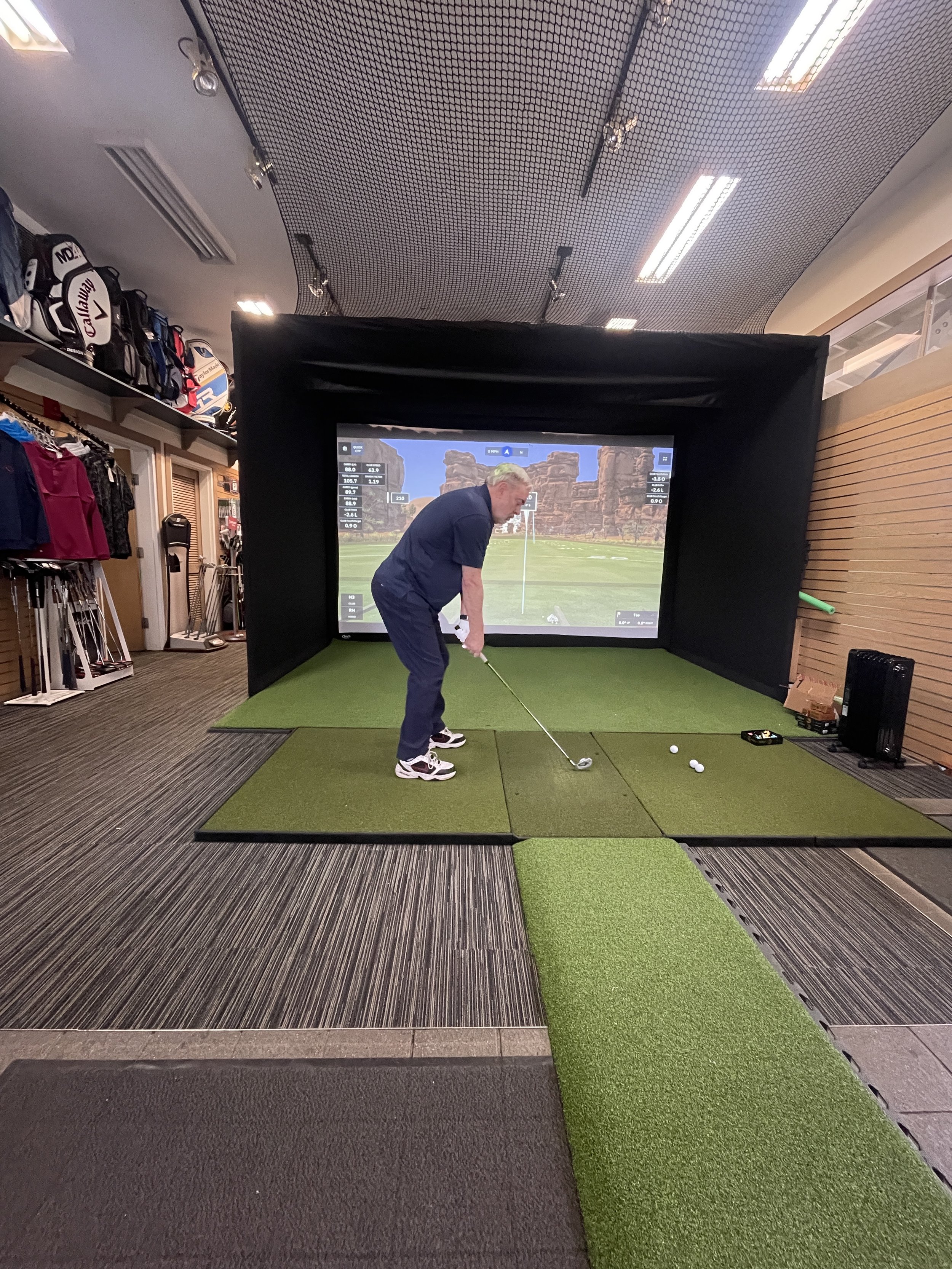 Man practicing golf indoors in a simulation room with a large digital screen displaying a golf course, wearing a navy shirt, blue pants, and white sneakers, preparing to hit a golf ball on artificial turf with golf clubs and balls around him.