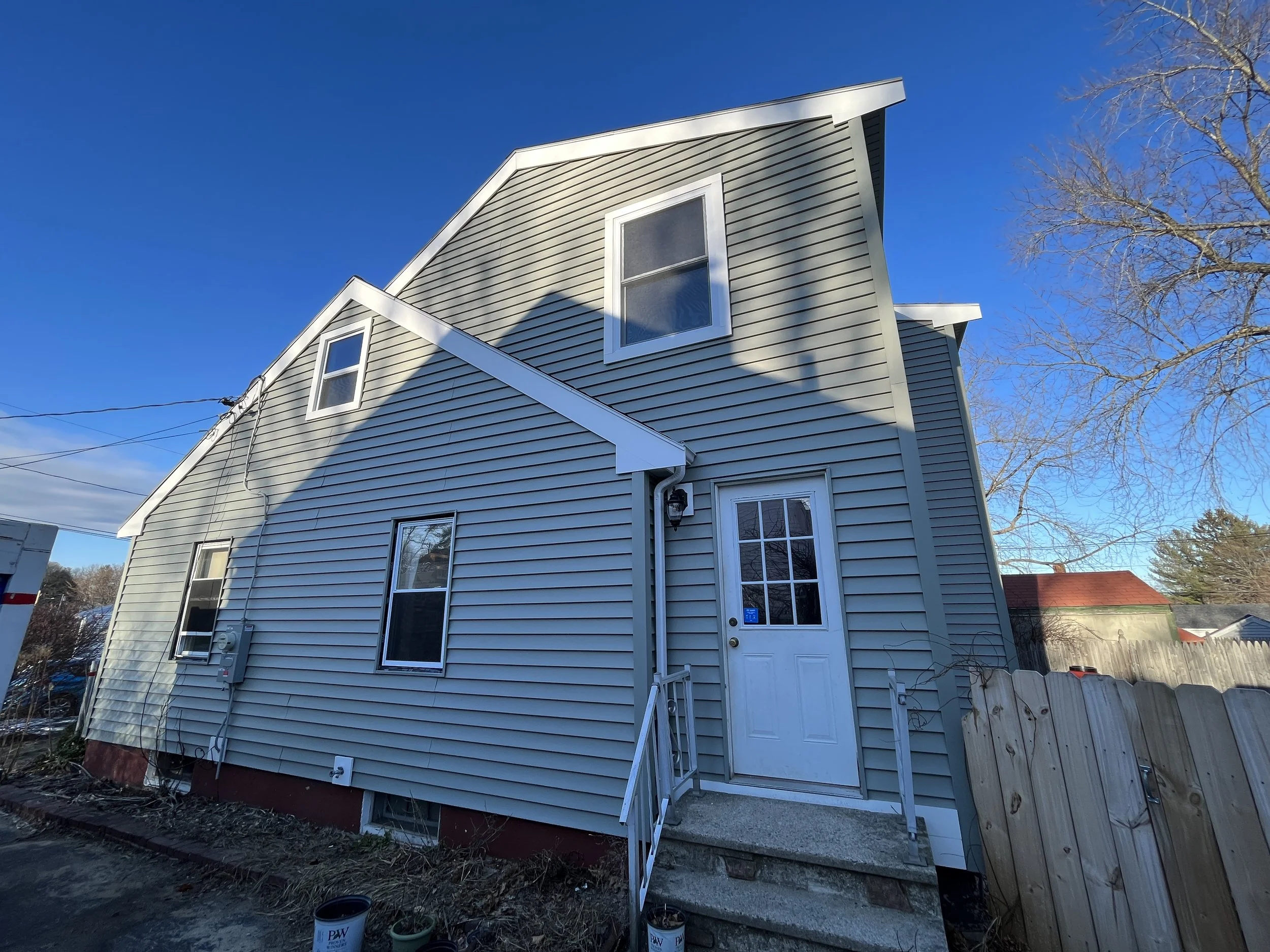 Side view of a two-story house with beige vinyl siding, white trim, and a small front porch with stairs, under a clear blue sky. A leafless tree is on the right side.