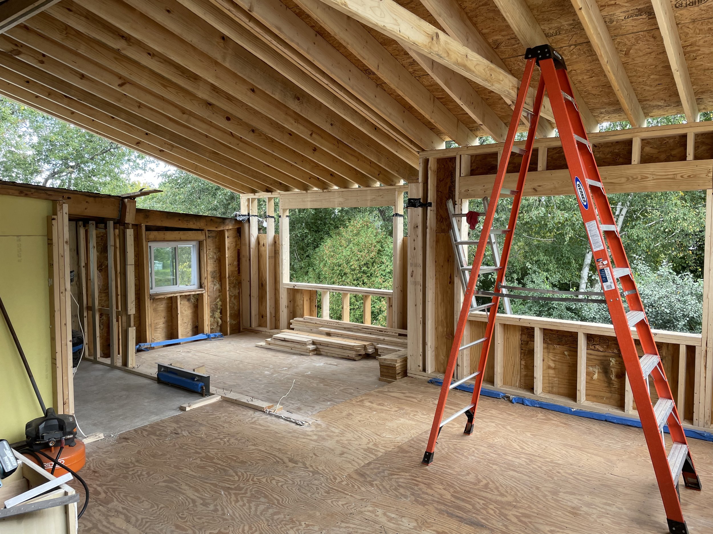 Interior of a house under construction, showing wooden framing, an extension ladder, a window opening, construction tools, and building materials.