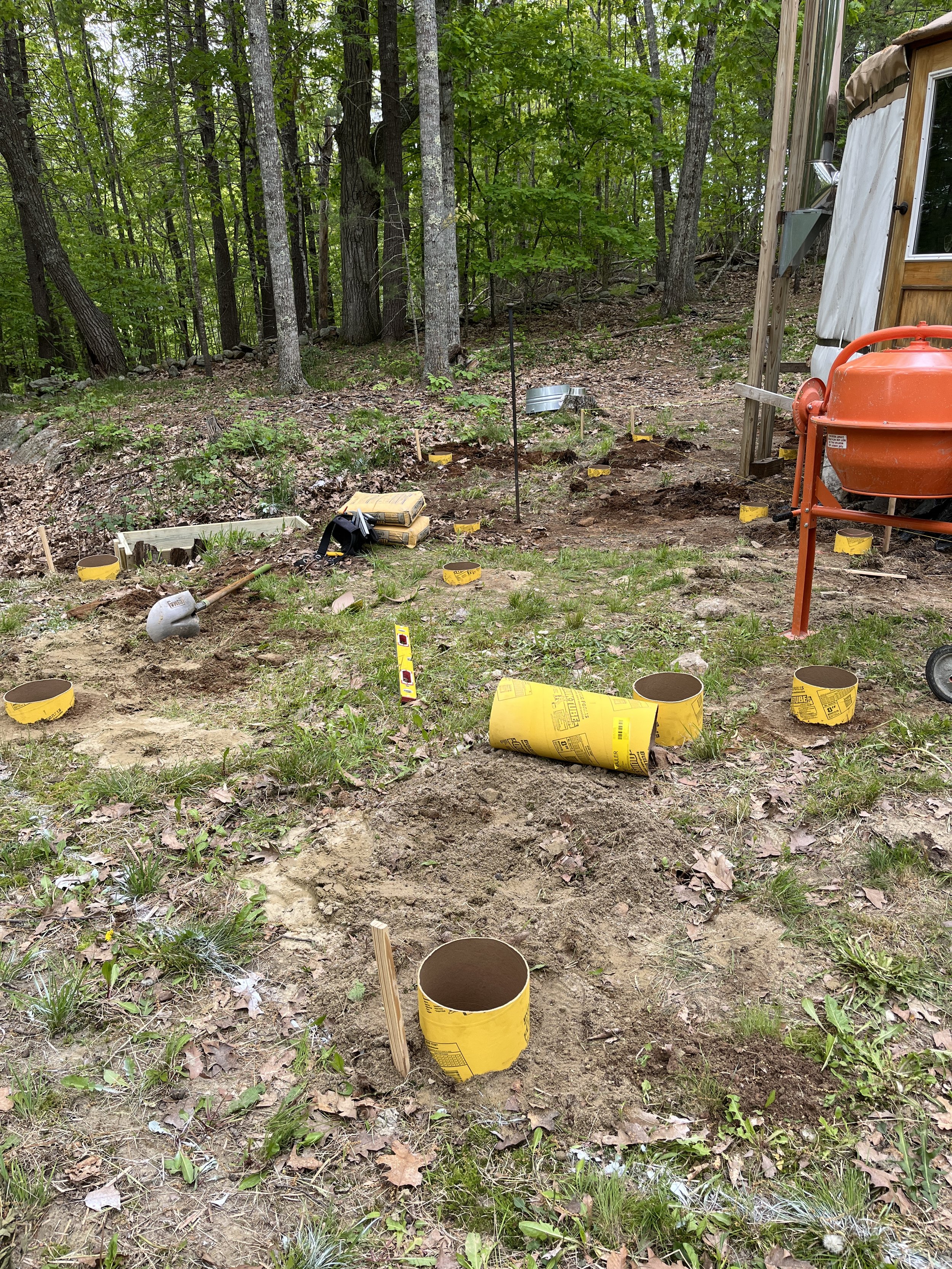 A construction site outdoors with drilled holes in the ground, yellow form tubes, and construction tools, surrounded by trees.