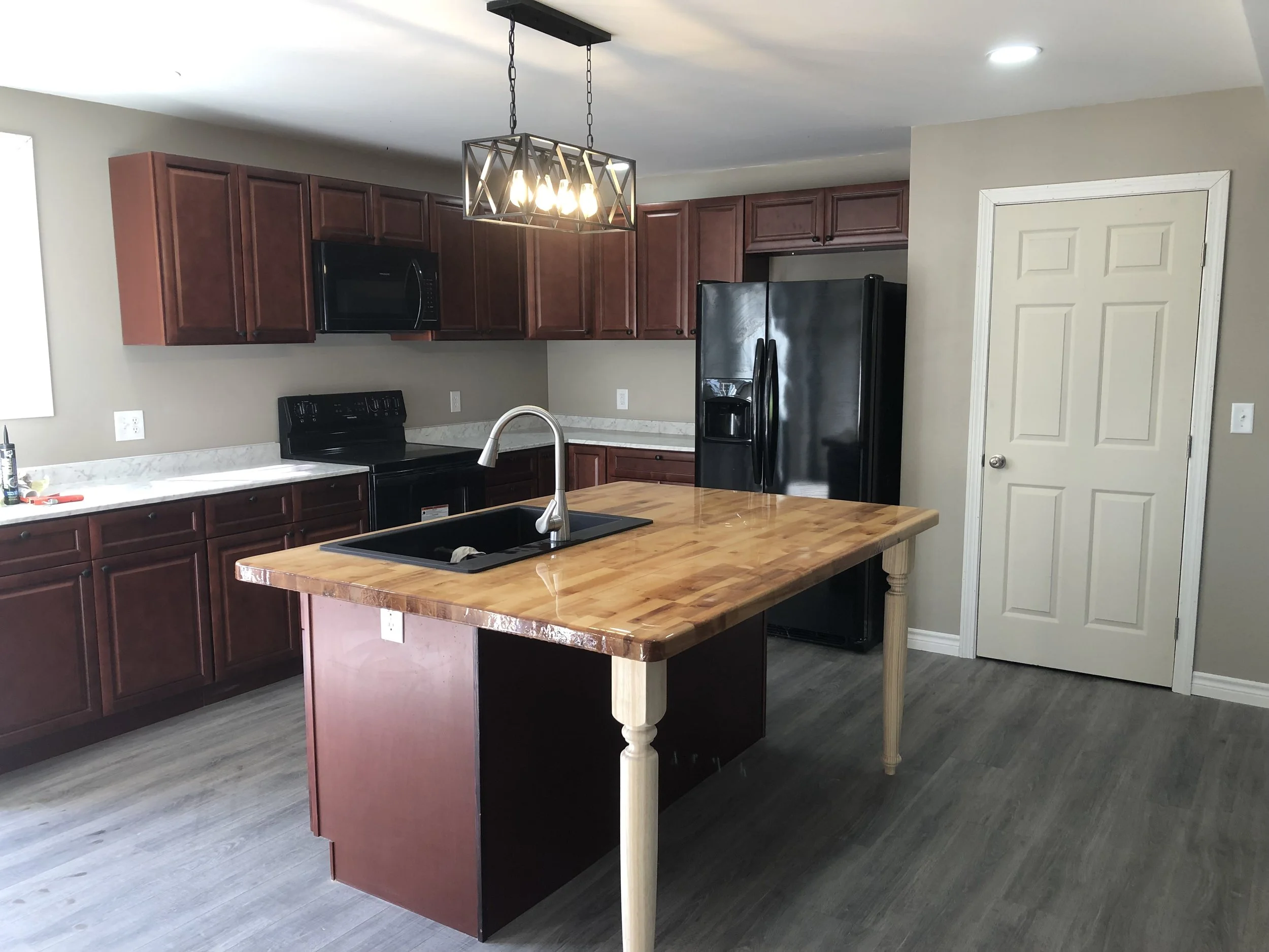 Modern kitchen with wooden cabinets, black appliances, a wooden central island with a sink, and a geometric light fixture overhead.