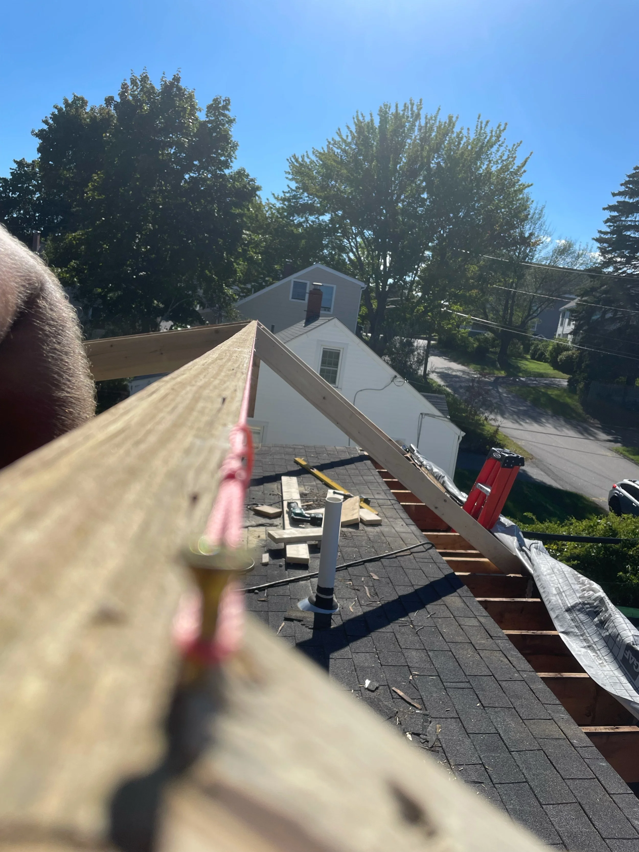View from a roof under construction showing a wooden beam, tools, shingles, and a suburban street with trees and houses in the background.