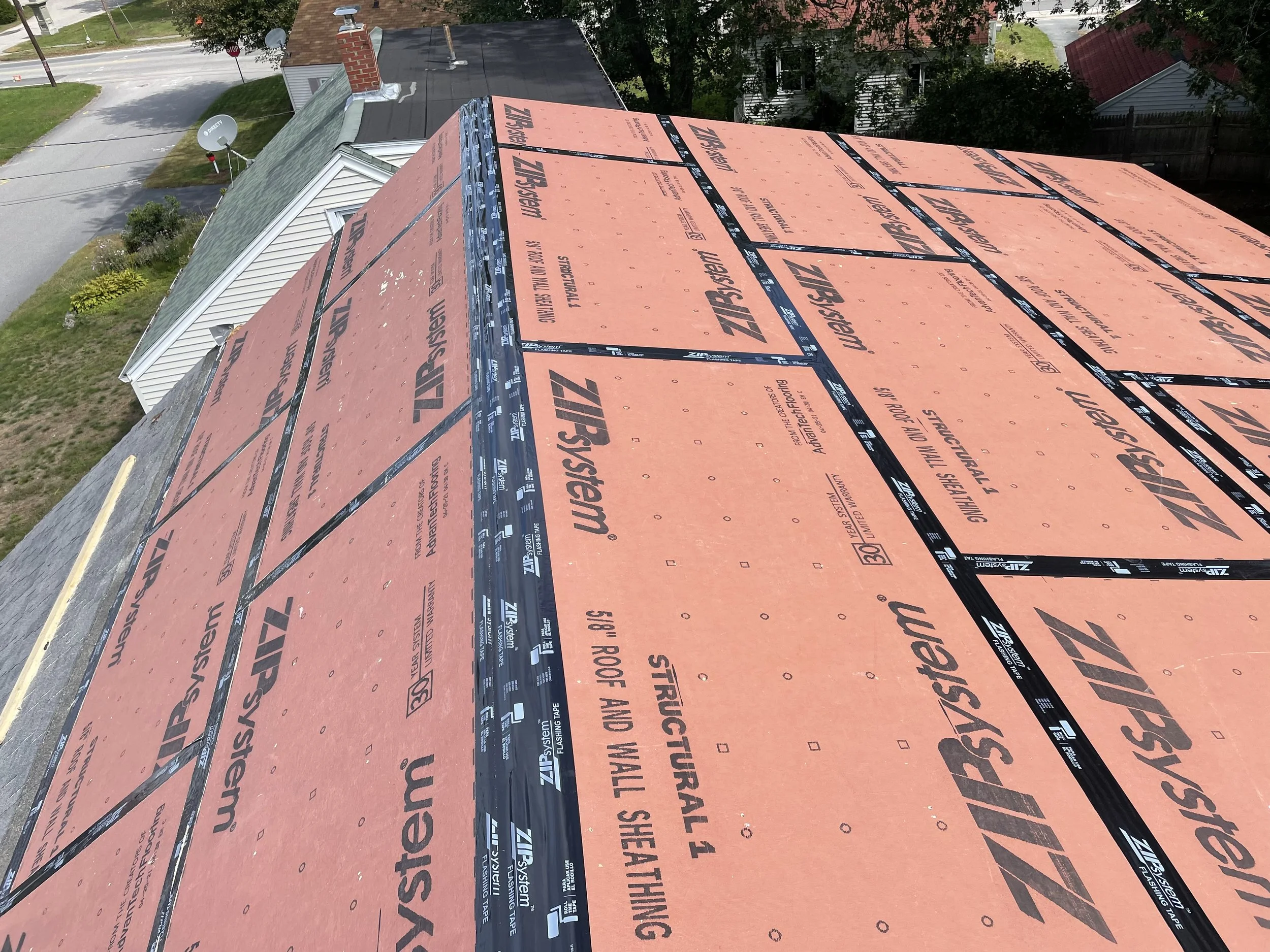 Roof under construction, covered with orange ZIP System sheathing panels and black tape, on a residential house with neighboring houses and trees visible.