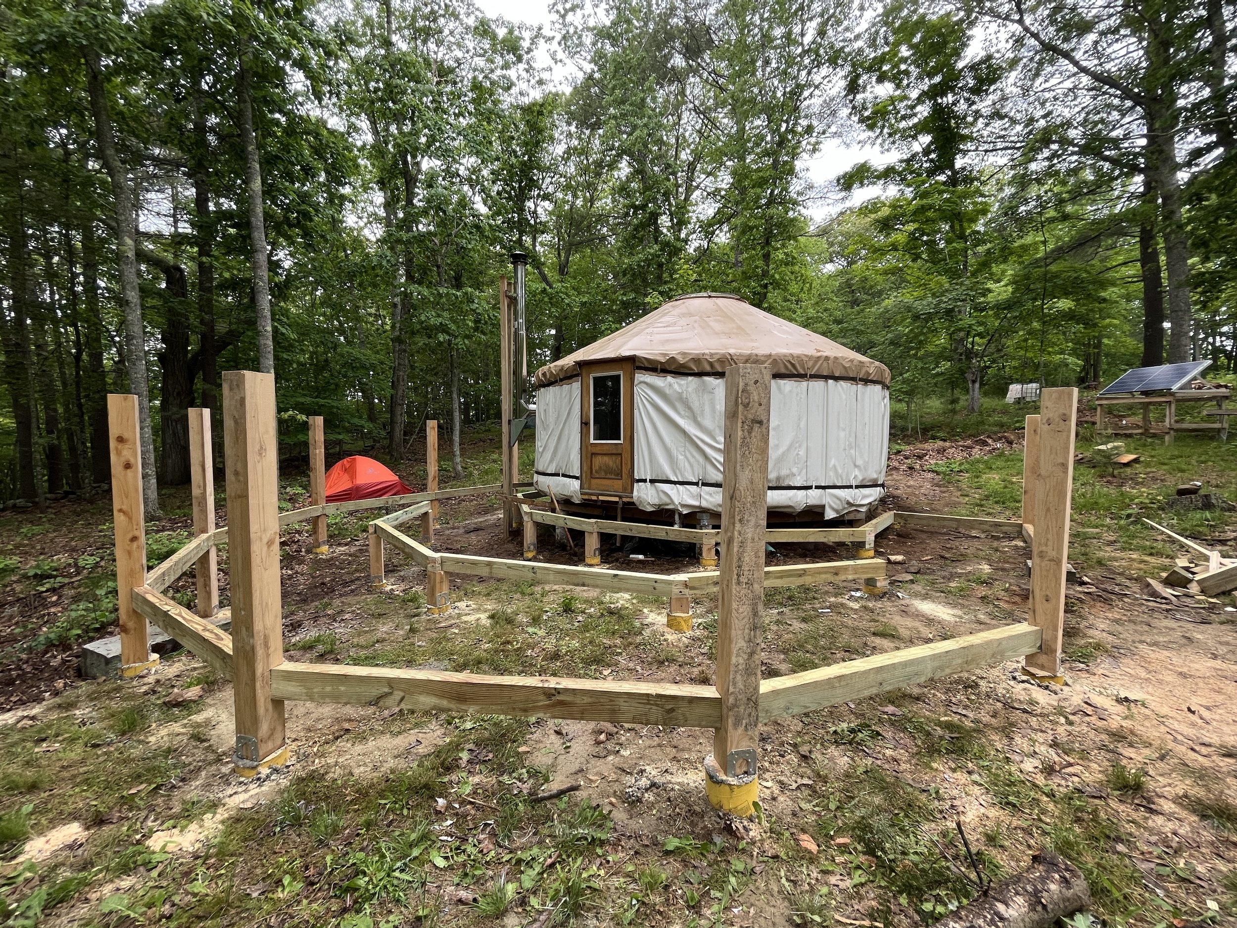 A yurt under construction in a wooded area, surrounded by a wooden fence, with a small tent and solar panel in the background.