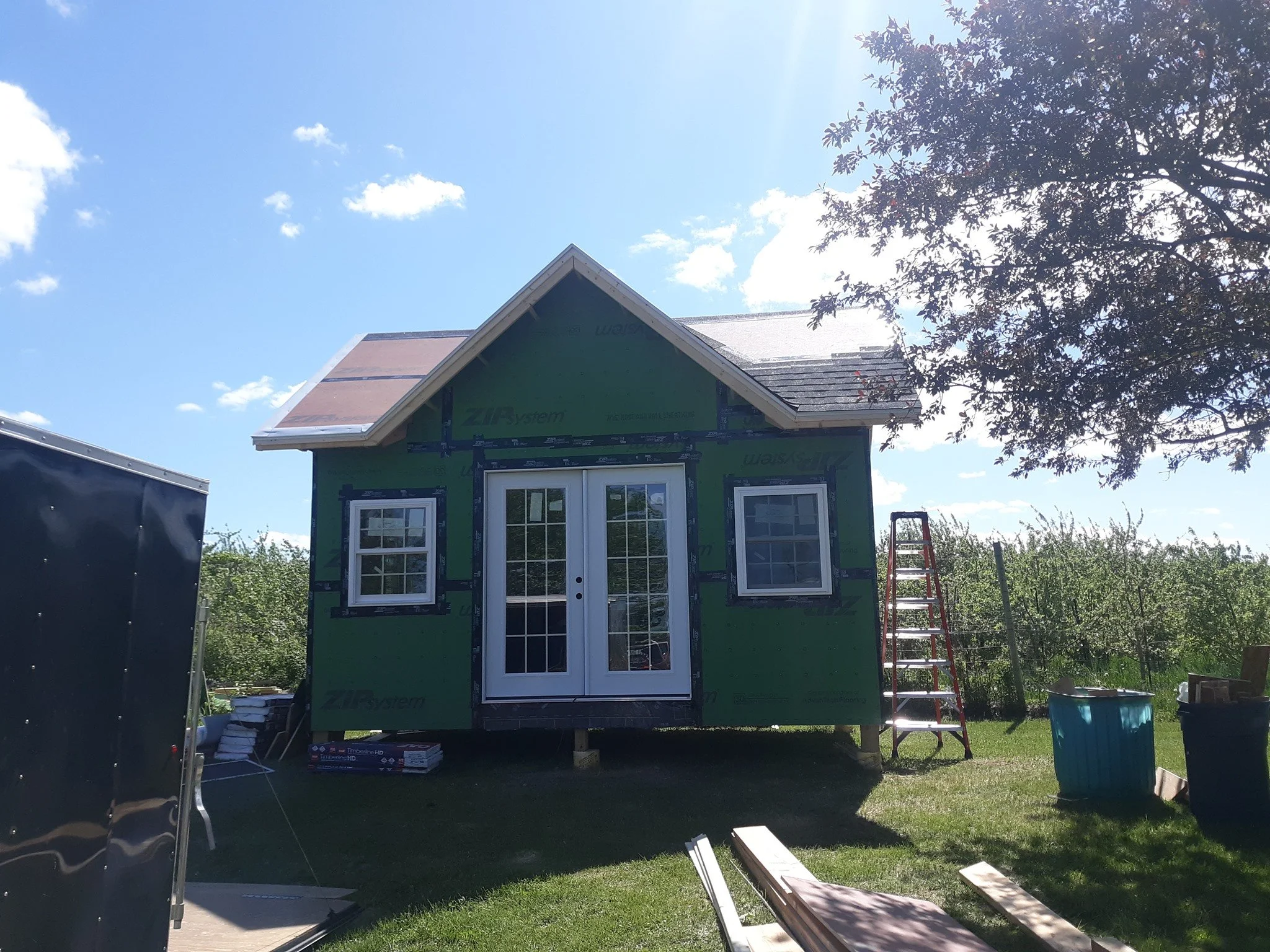 A small house under construction with green sheathing, a double door, two windows, and a sloped roof, on a grassy area with construction materials and tools around.