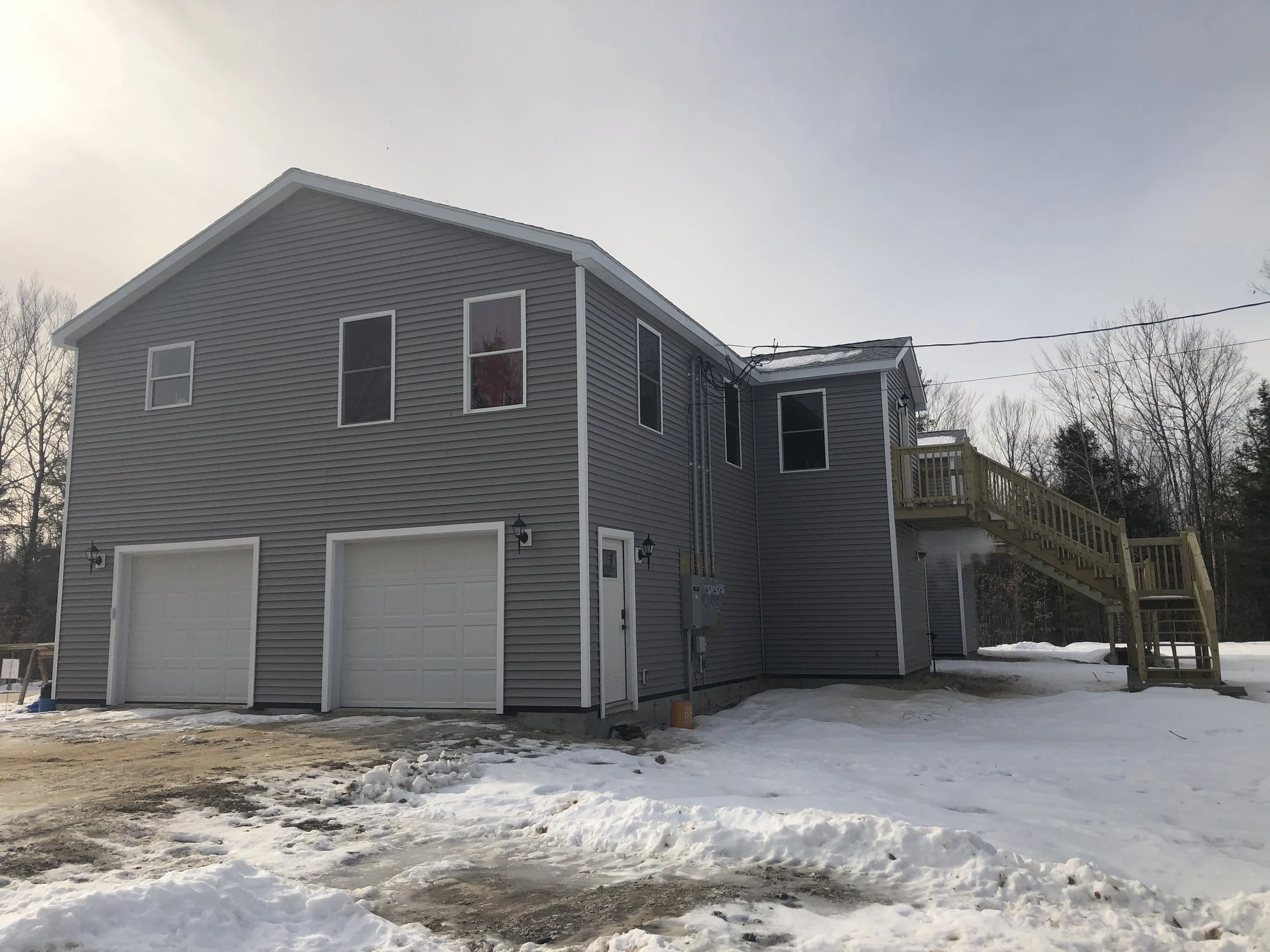 A two-story gray house with vinyl siding, two garage doors, a side staircase, and snow on the ground.