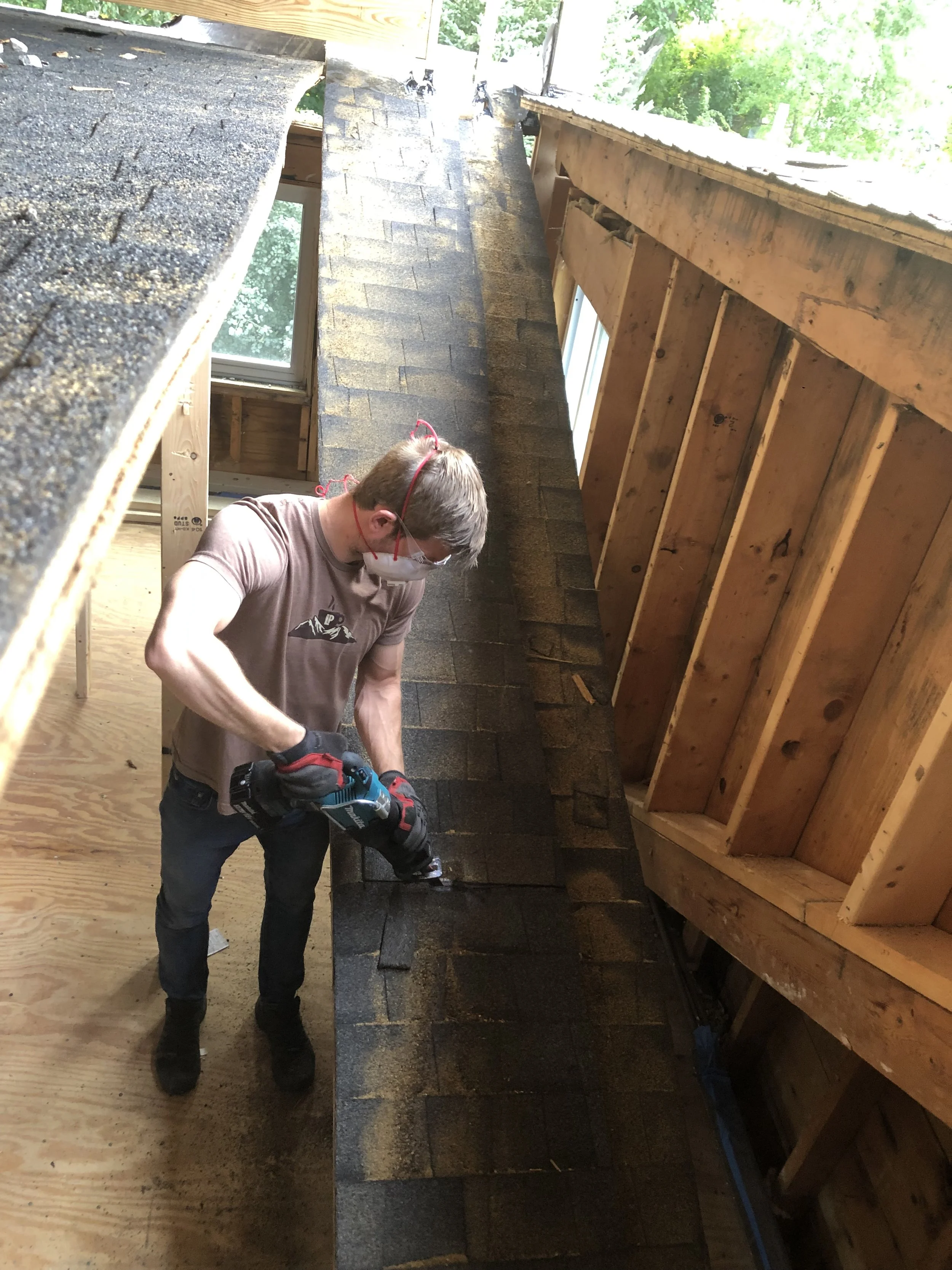 A person installing or repairing shingles on a roof, wearing a face mask, glasses, gloves, and using a cordless drill. The roof has new and old shingles, with exposed wooden framing and windows visible.