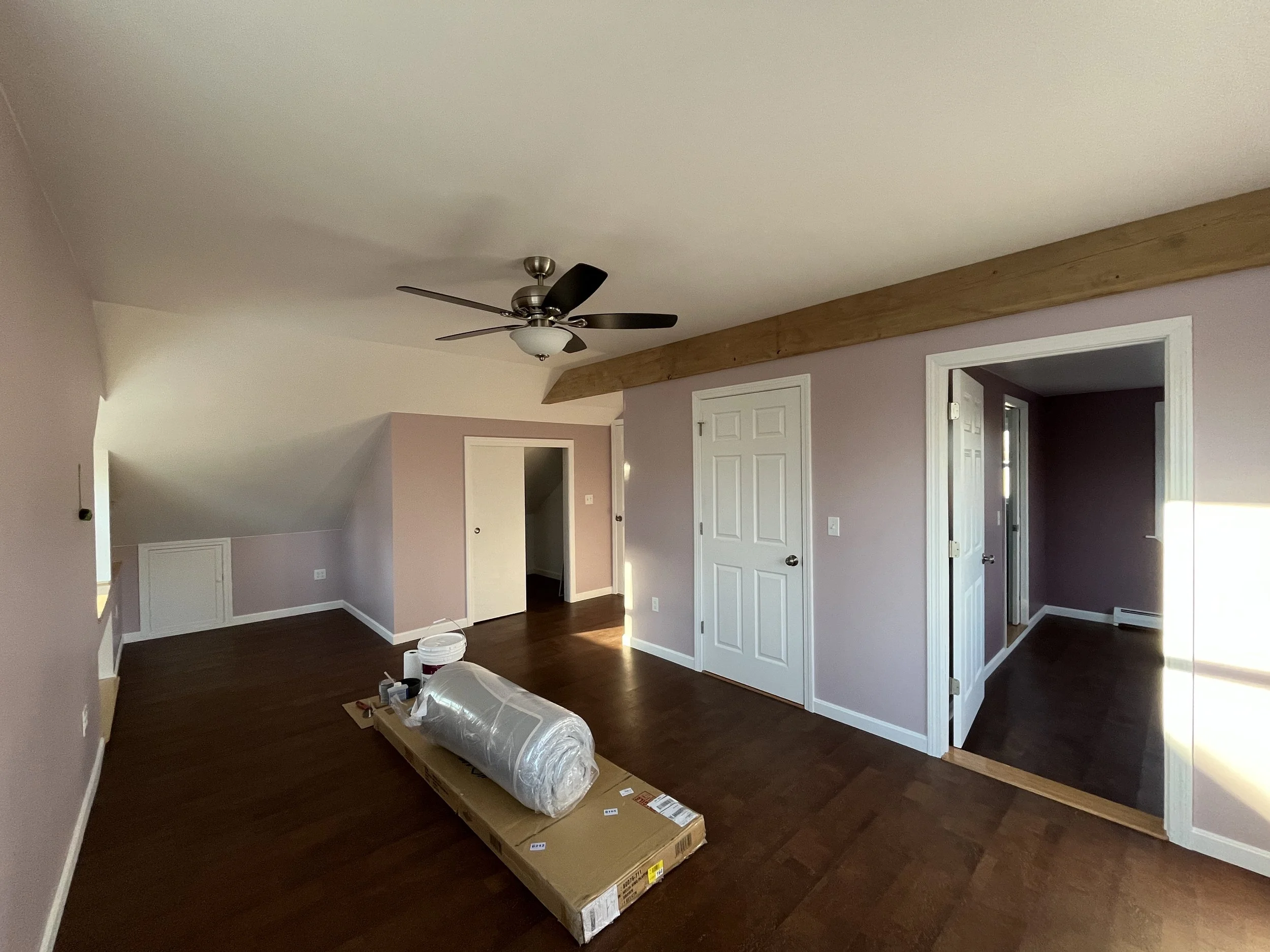 Empty living room with pink walls, dark hardwood floors, a ceiling fan, and a wooden beam near the ceiling. There are two doorways, one leading to a small room and another to a hallway, and a window on the right side. Artwork and furniture are not pr