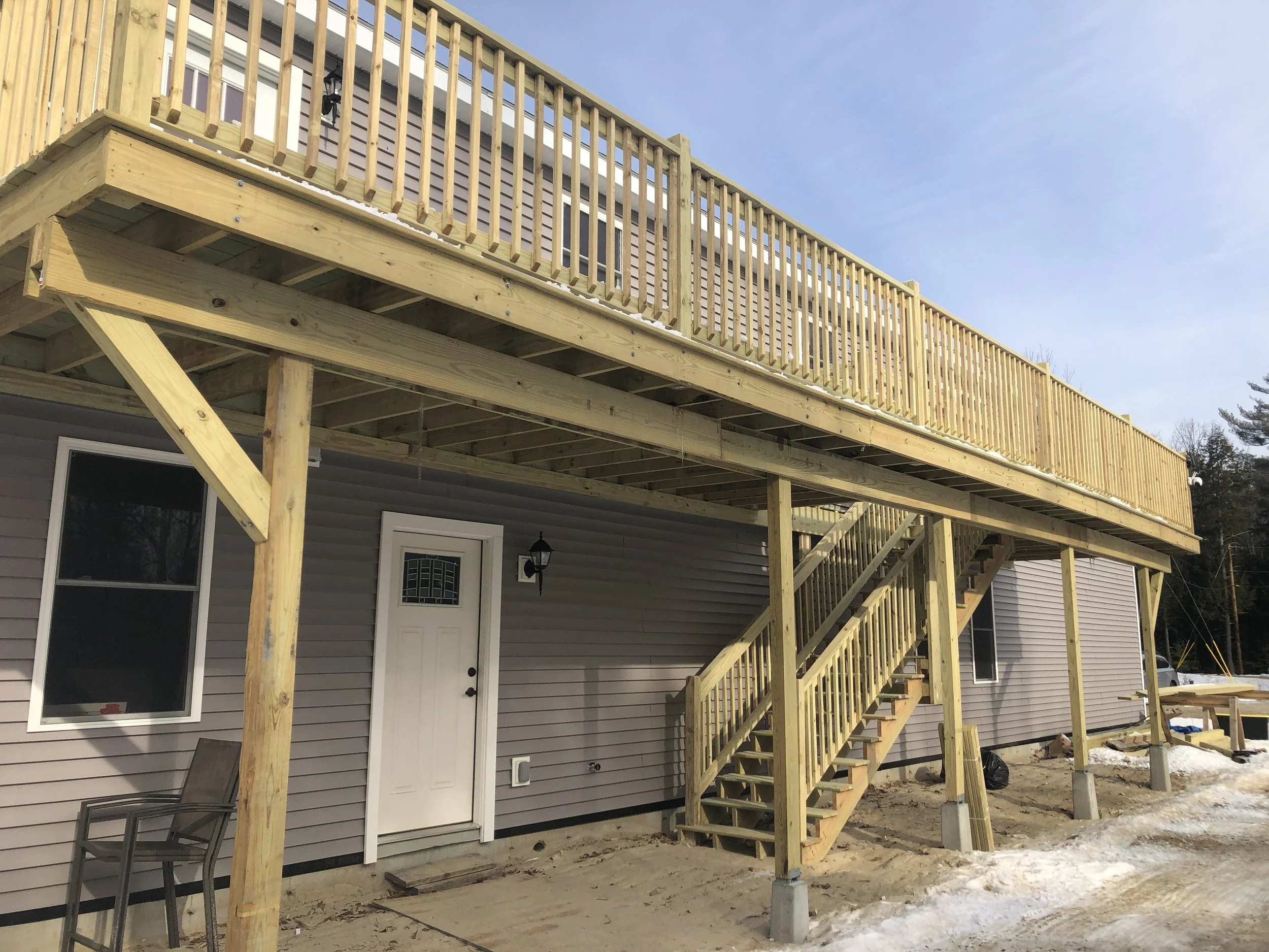 New wooden deck with stairs under construction attached to the back of a house, with support posts anchored in concrete, in a yard with snow patches.