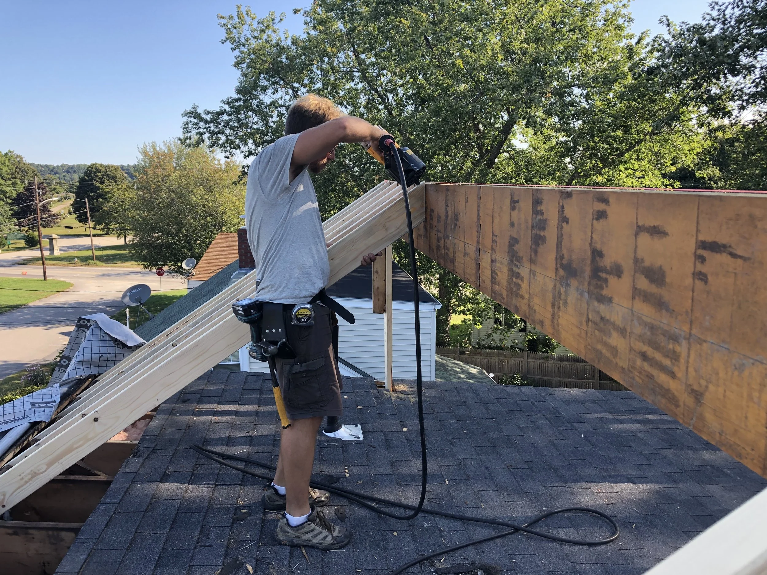 A person working on the roof of a house, using a power drill on a wooden beam under a tree, with a neighborhood in the background.