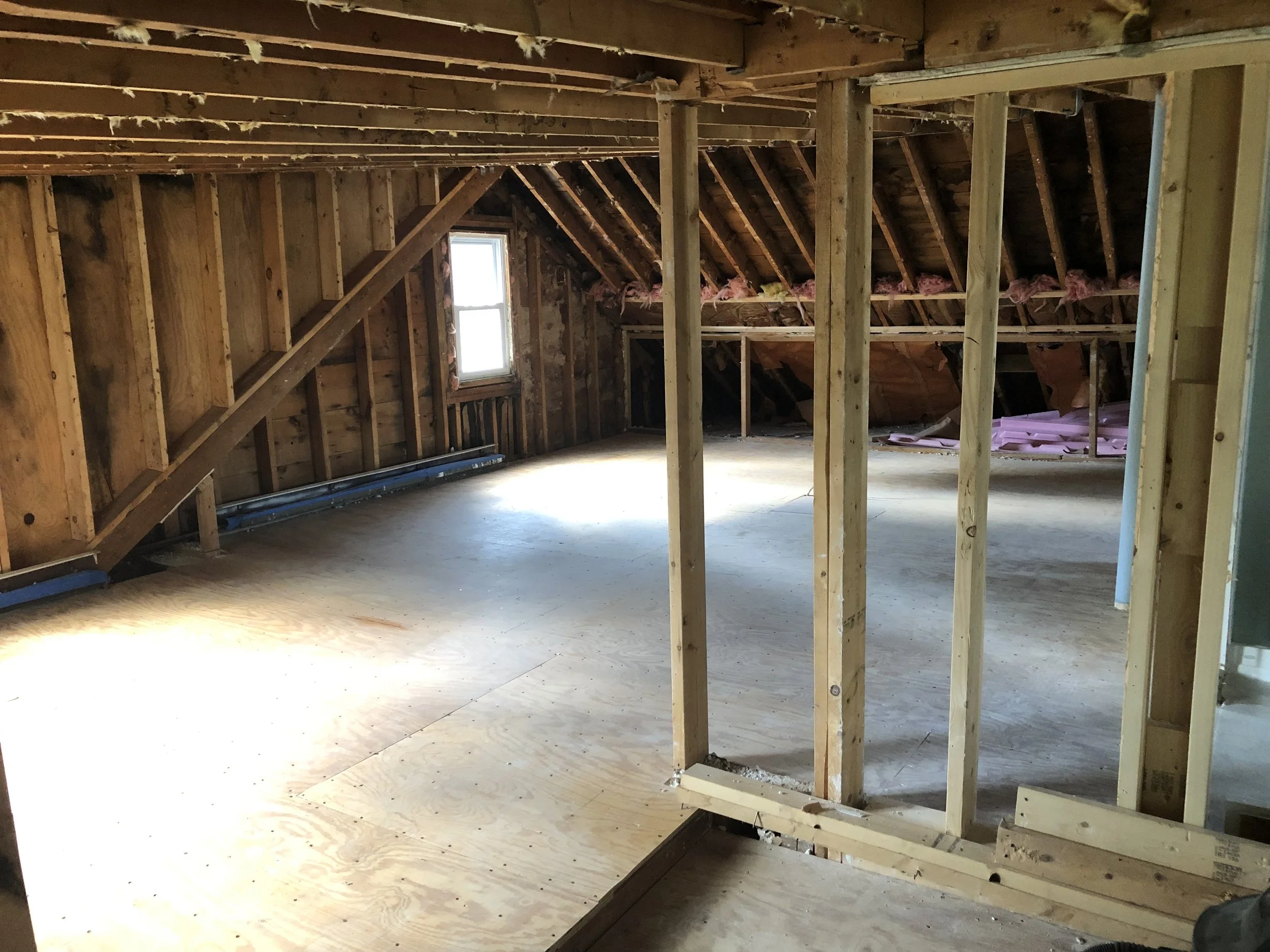 Interior of an unfinished attic with exposed wooden studs, rafters, and insulation along the edges, with a small window on the left wall letting in natural light.