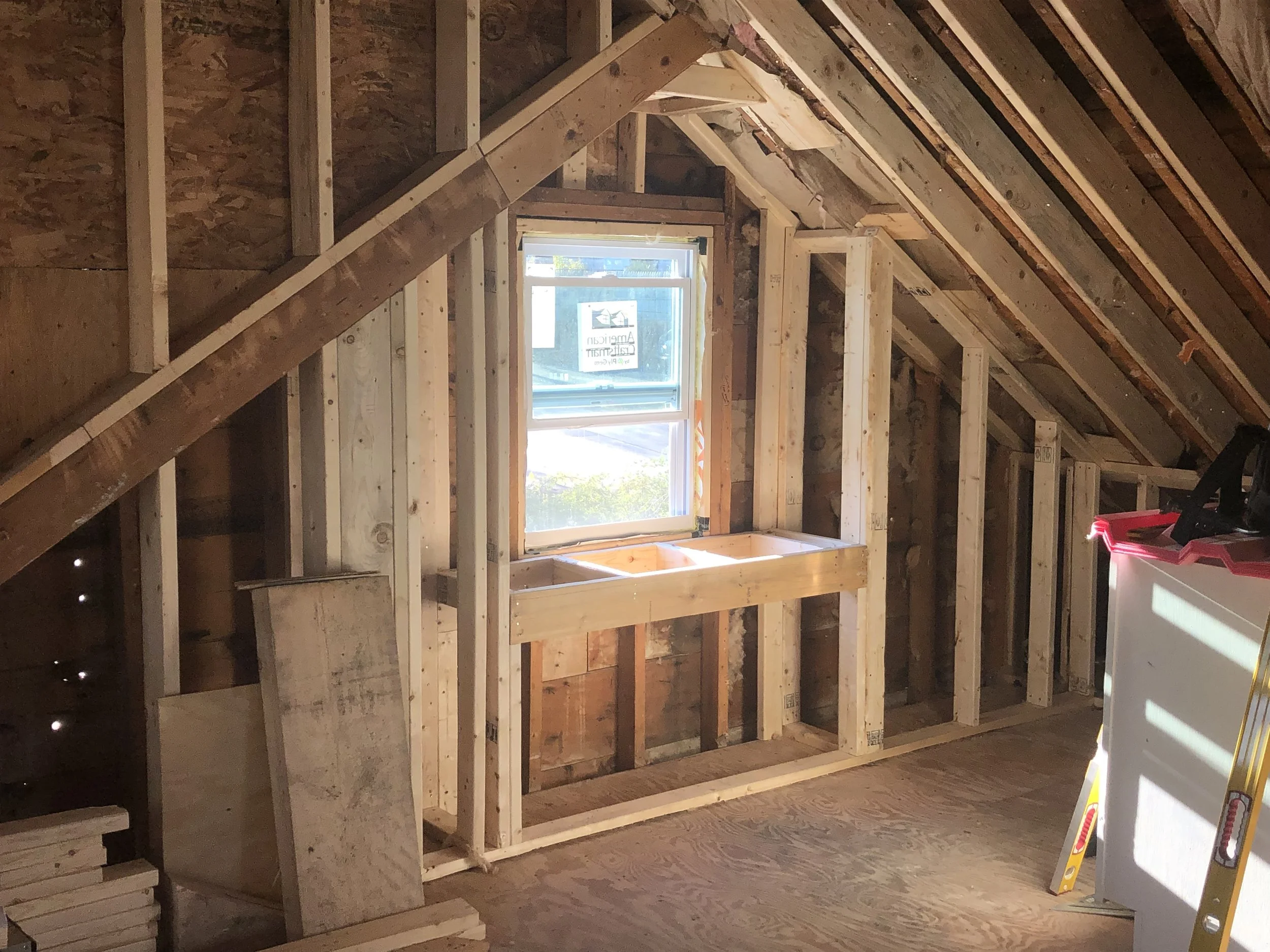 Interior of an attic under construction with exposed wooden framing, a window, and some construction tools and materials.