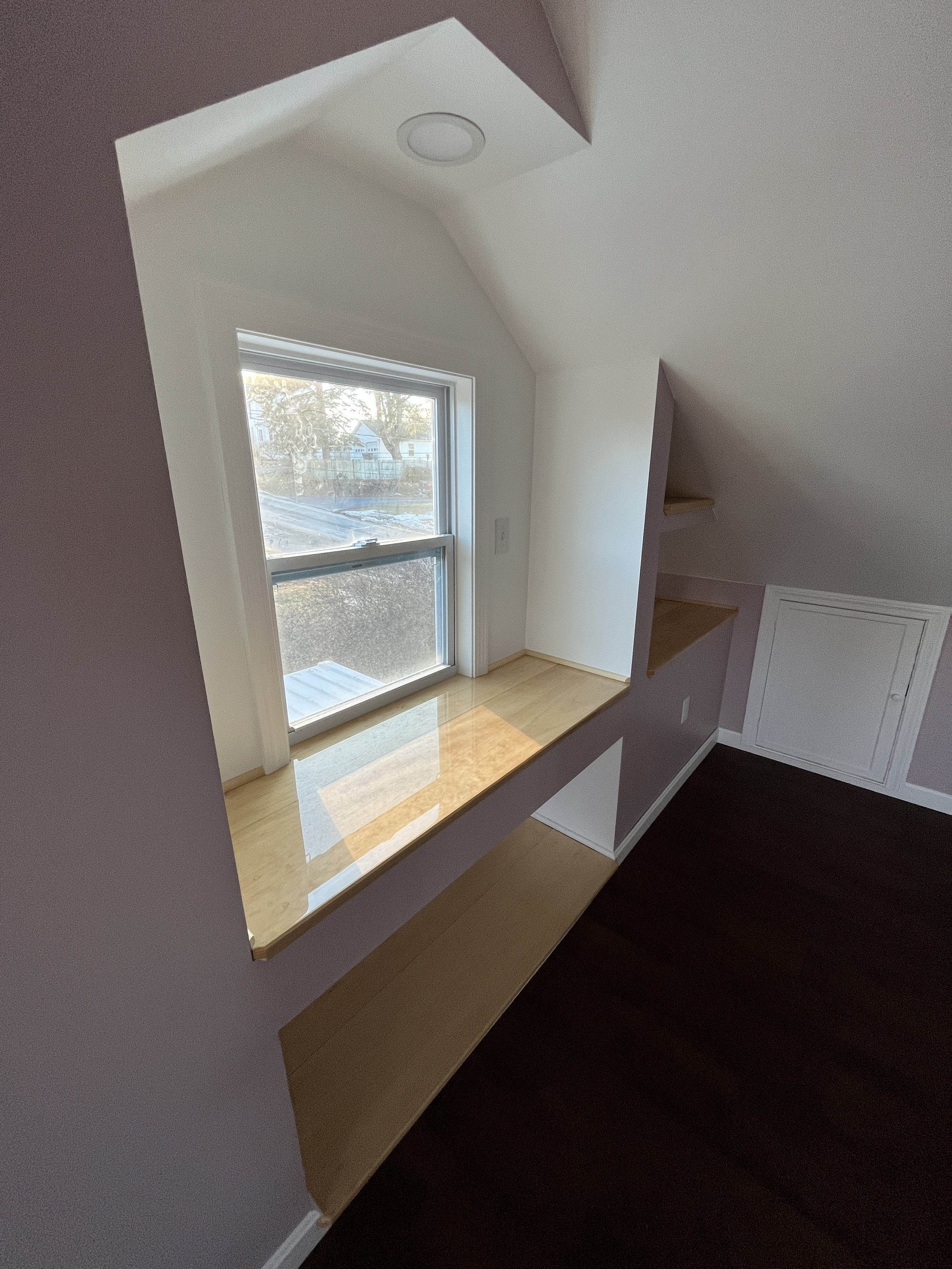 A small nook with a window, a wooden window seat, and shelves in a room with light-colored walls and dark wood flooring.