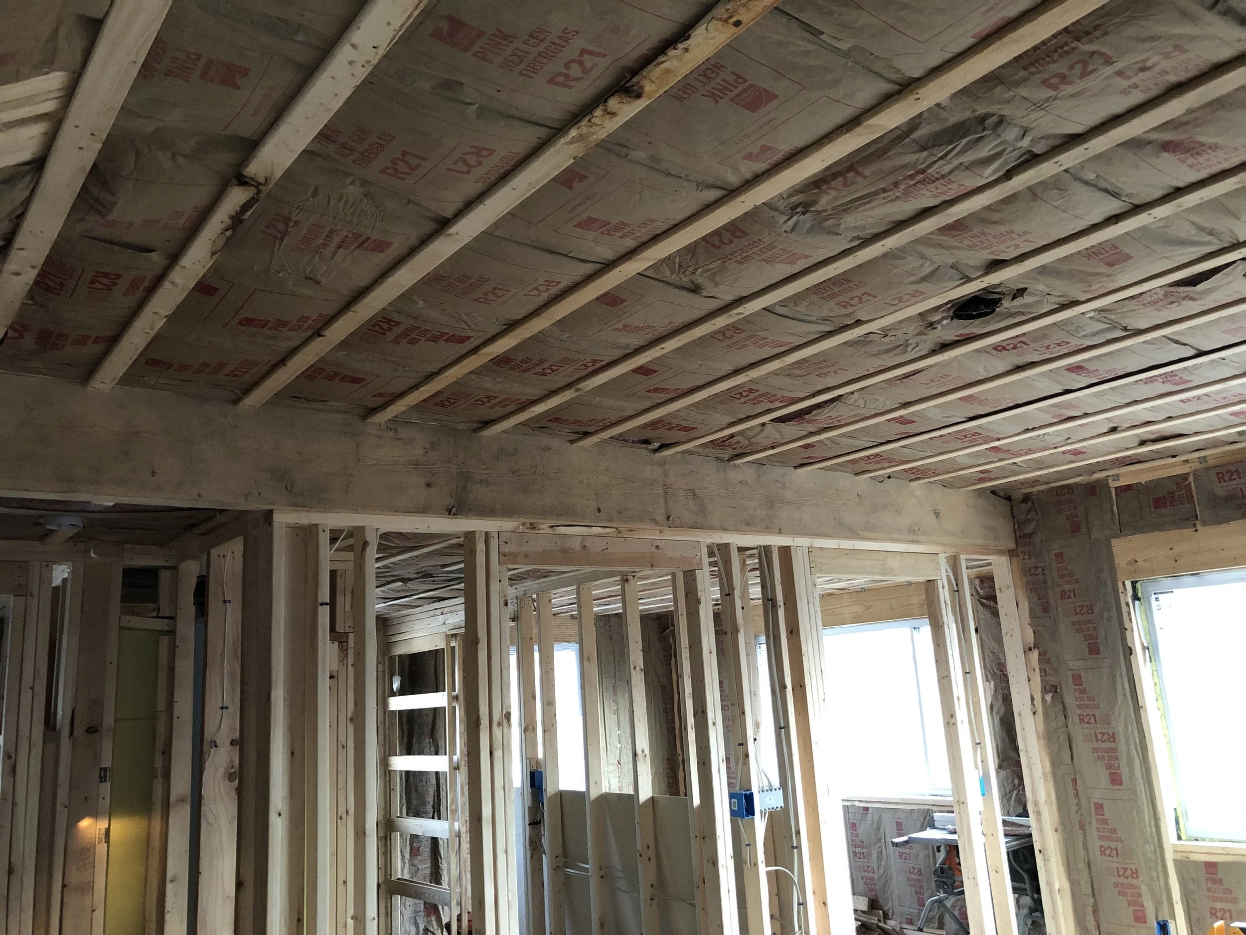 Interior view of a house under construction with exposed wood framing, insulation in the walls and ceiling, and windows.