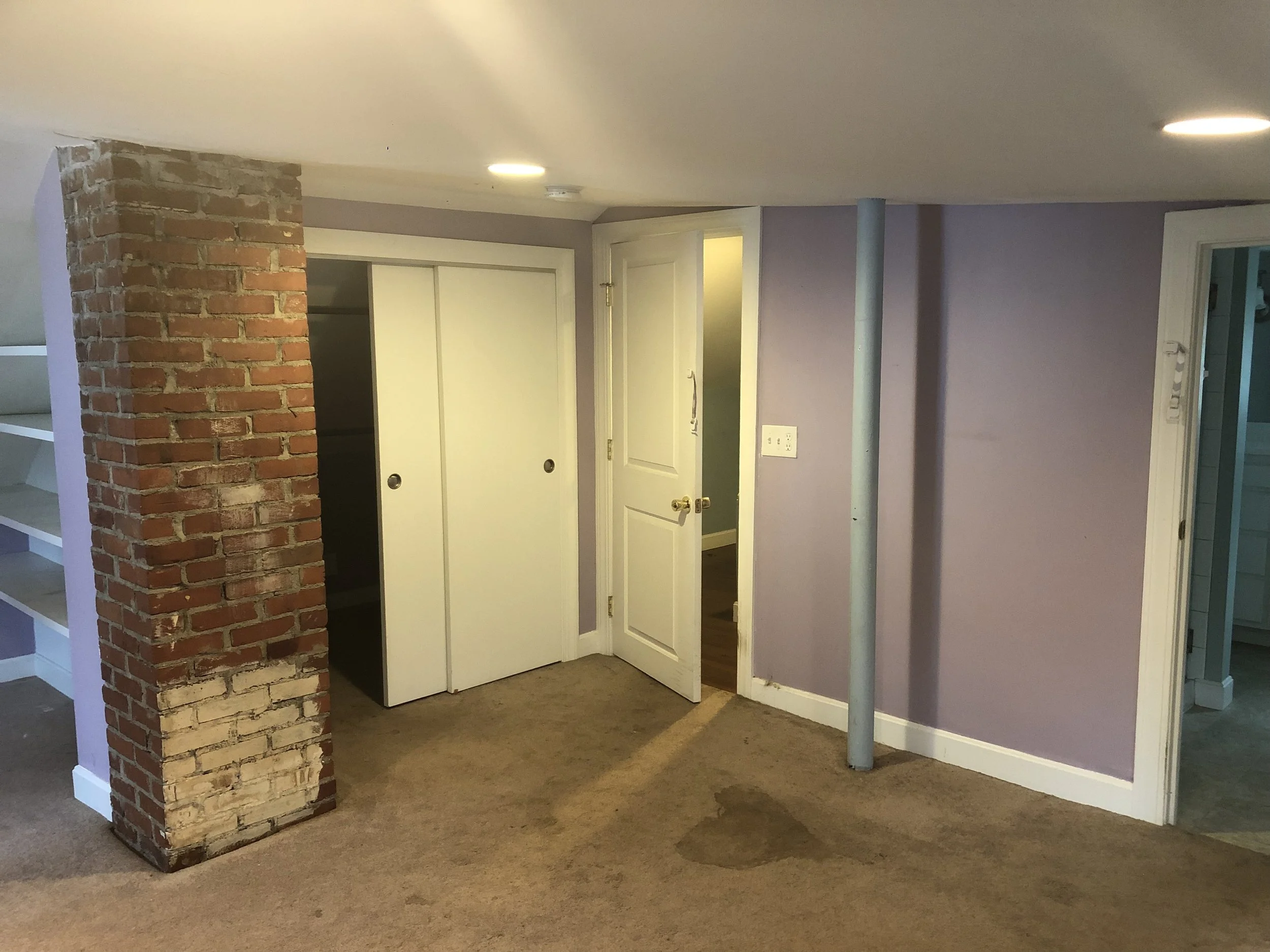 Basement room with purple walls, brown carpet with stains, brick pillar, white closet doors, open door to hallway, and pipe running vertically along wall.
