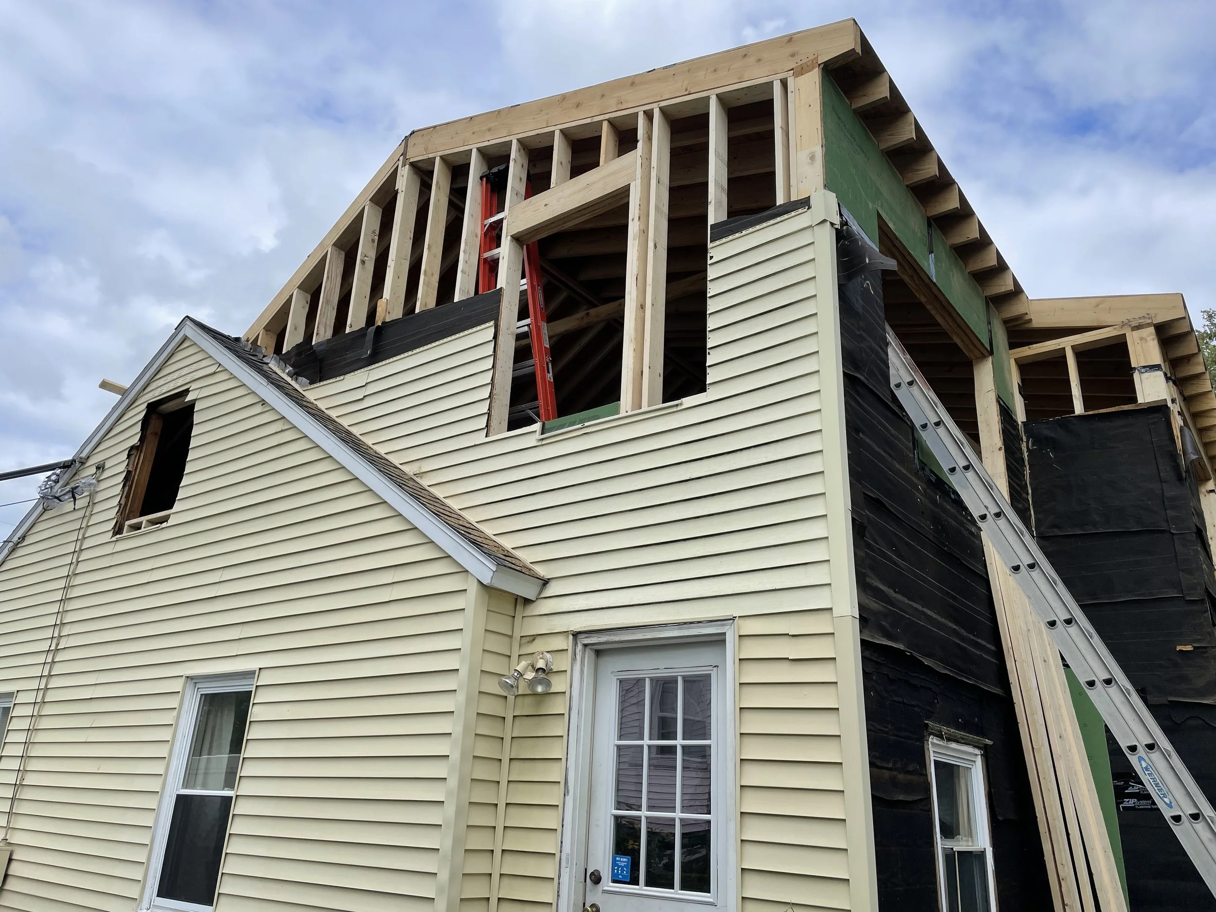 A two-story house under construction with missing siding on the upper level, a red ladder inside the open area, and black weatherproofing material on parts of the exterior wall. The lower level has beige vinyl siding, with a window and a door visible