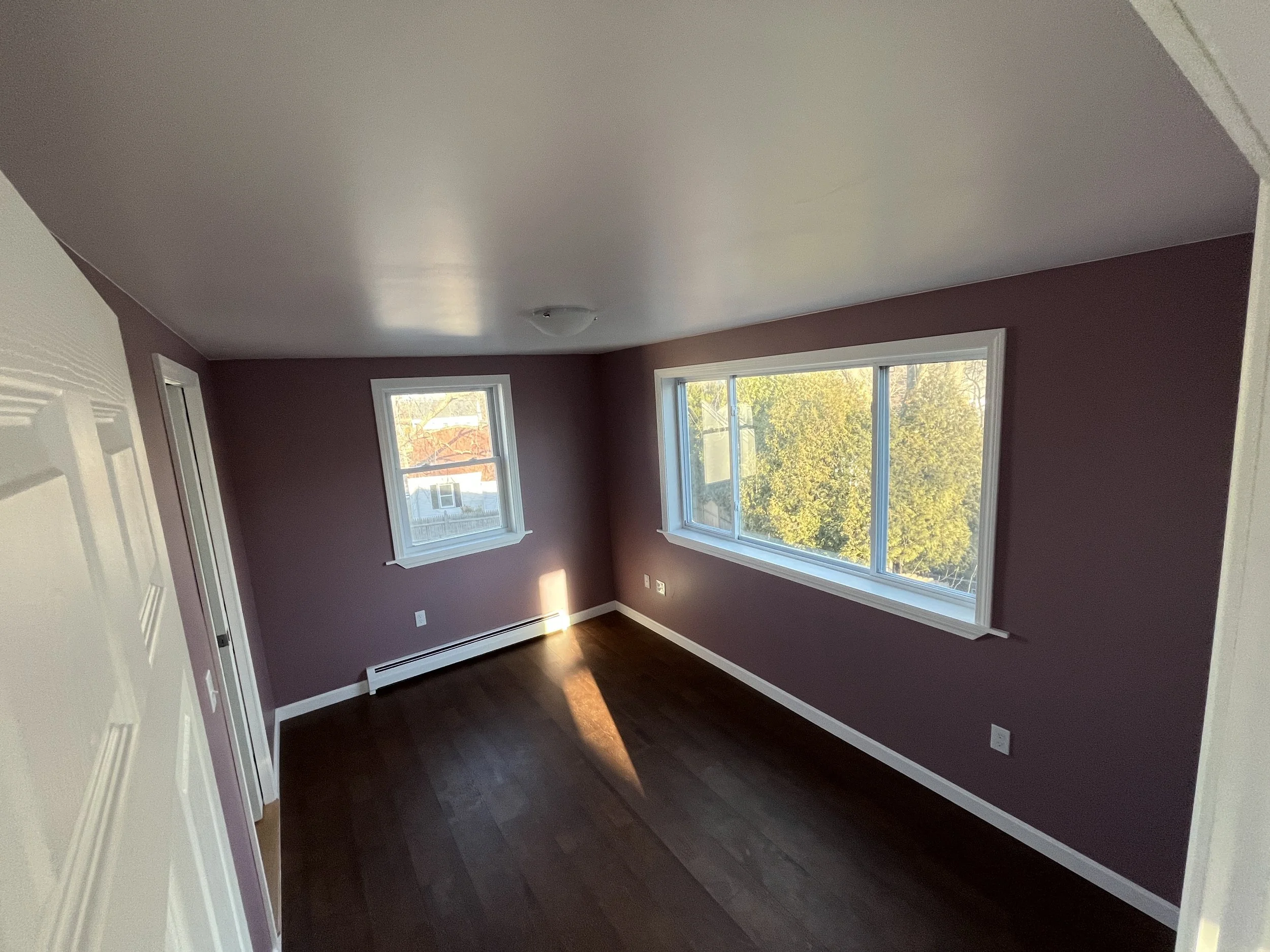 Empty room with purple walls, large window, small window, hardwood floor, and white baseboards and trim, with sunlight shining through.