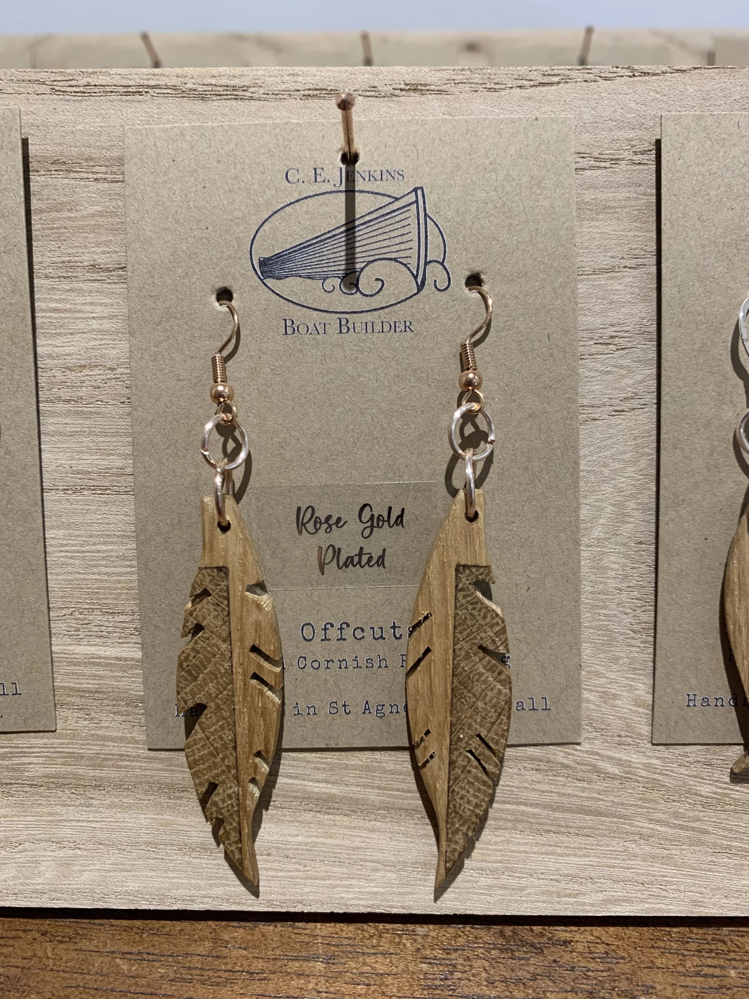 Wooden feather-shaped earrings with carved details, attached to a display card labeled 'C. E. Jenkins Boat Builder' and 'Rose Gold Plated', hanging on a wooden display.
