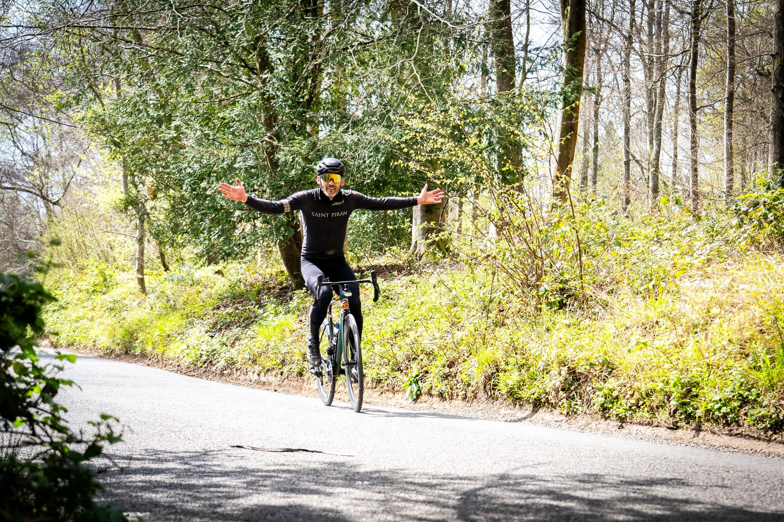 A man in cycling gear riding a bike on a forested road with arms outstretched and smiling.
