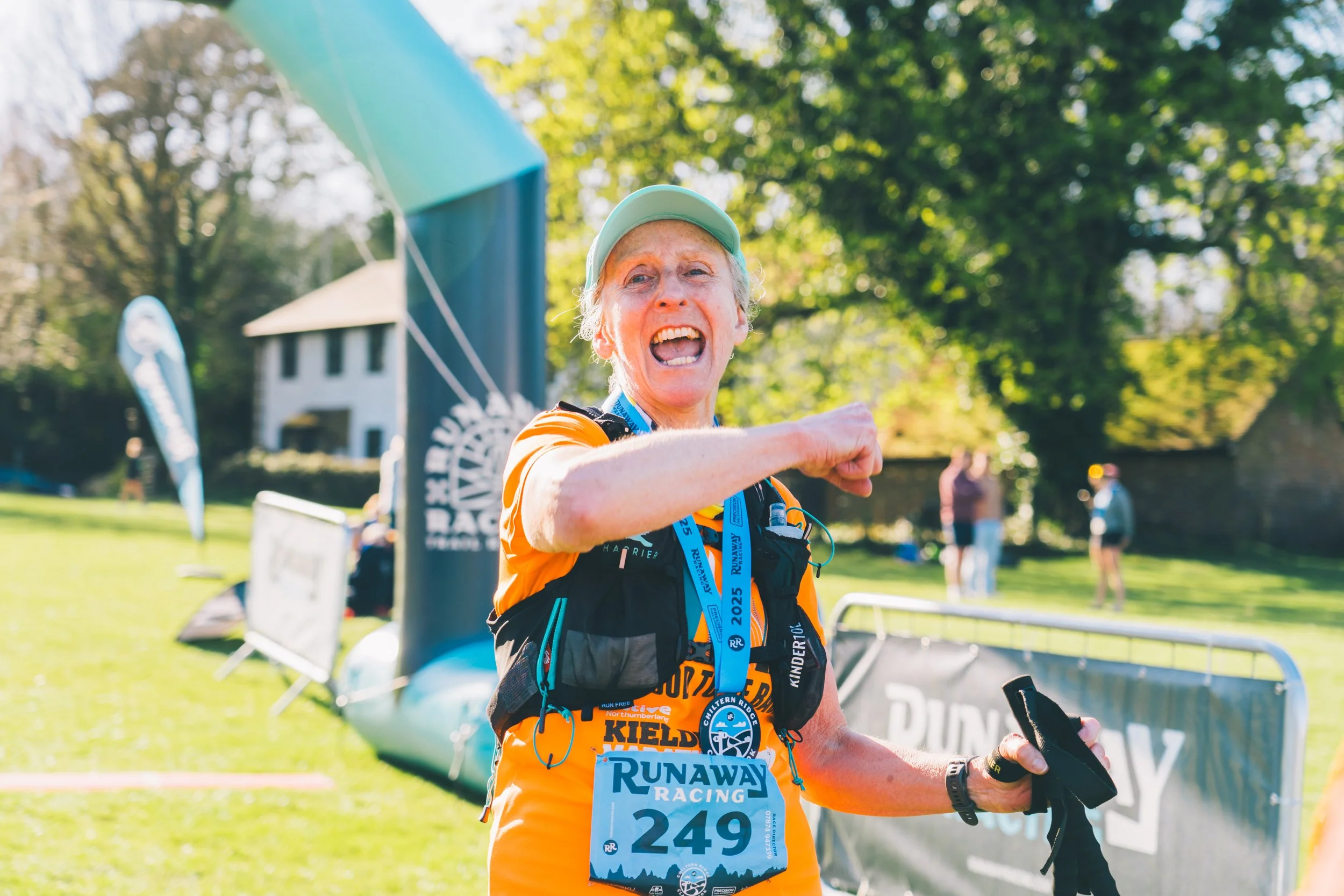 An elderly woman in running gear celebrating at the finish line of a race, smiling with her arm raised, wearing a medal and race bib number 249, outdoors with green trees and a building in the background.
