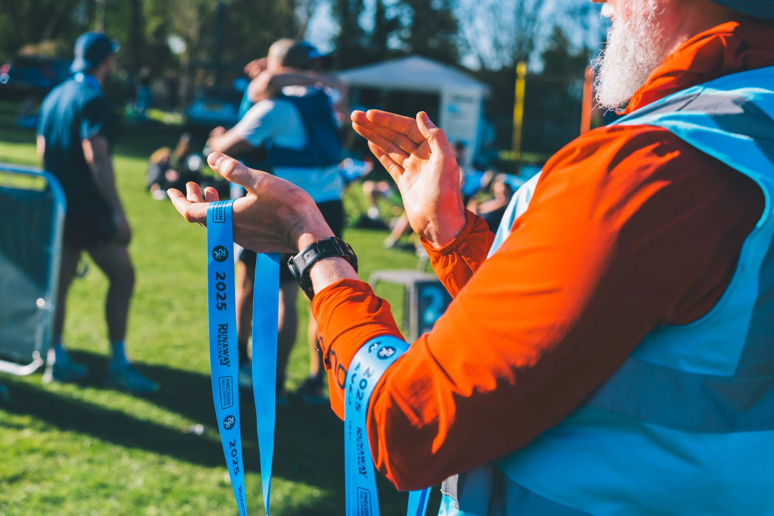 A man in a bright orange jacket is holding blue race medals at an outdoor event, with a grassy field and people in the background.