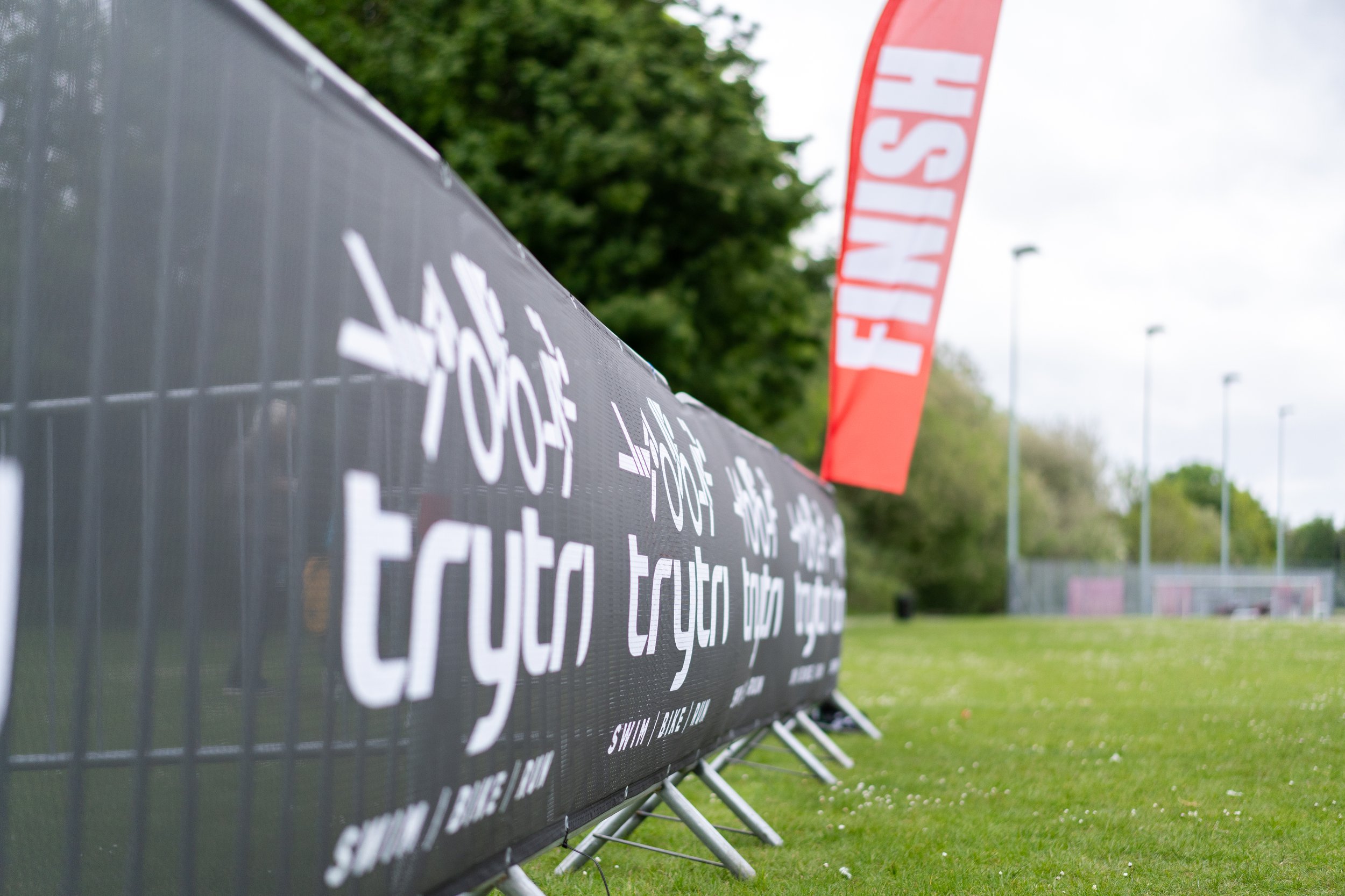 Large black advertisement banner with white text along a grassy sports field. Red flags with white FISH logos in the background. Trees and a cloudy sky overhead.