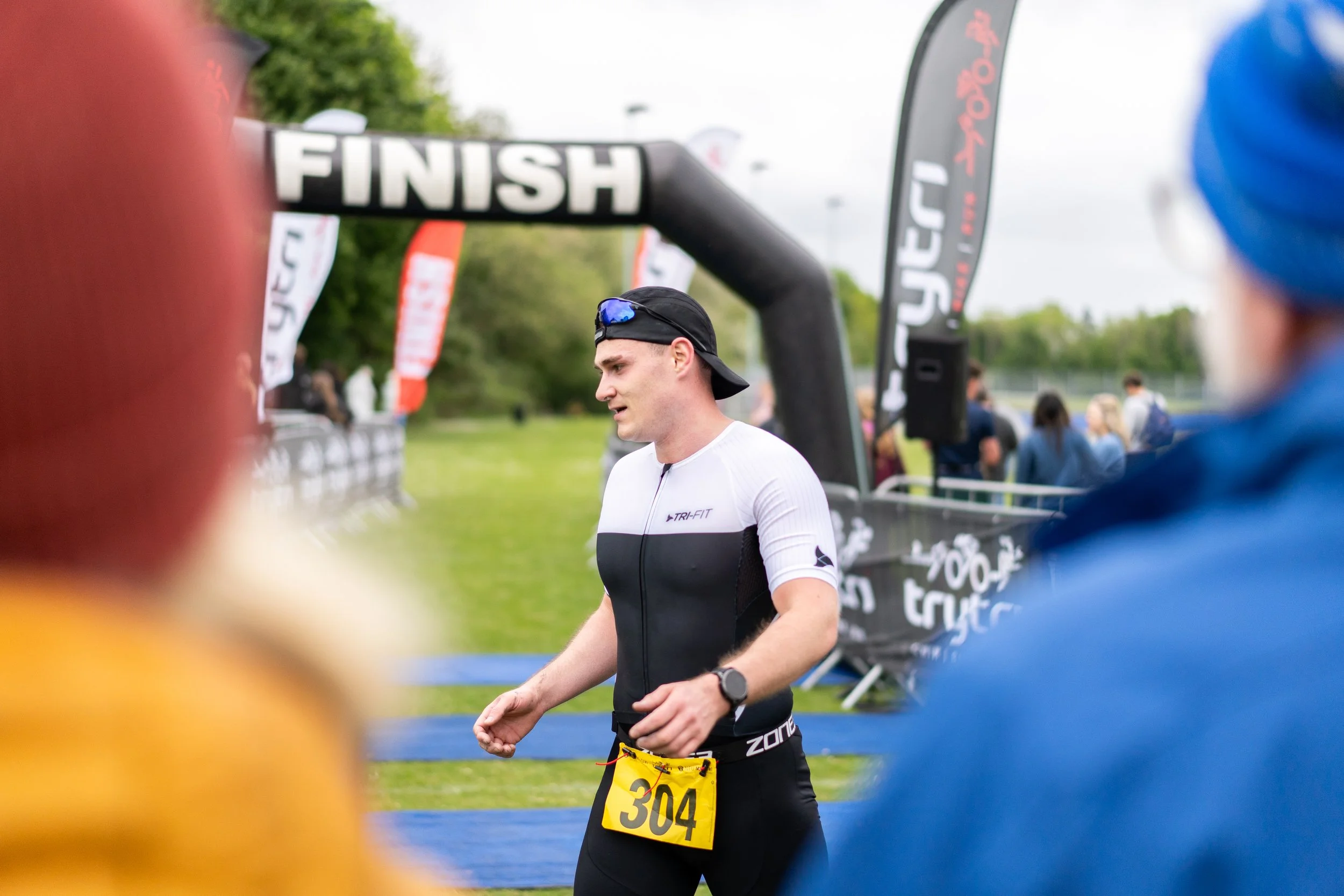Male marathon runner wearing a black cap, white and black athletic shirt, and a yellow race bib with the number 304, crossing the finish line with people and flags in the background.