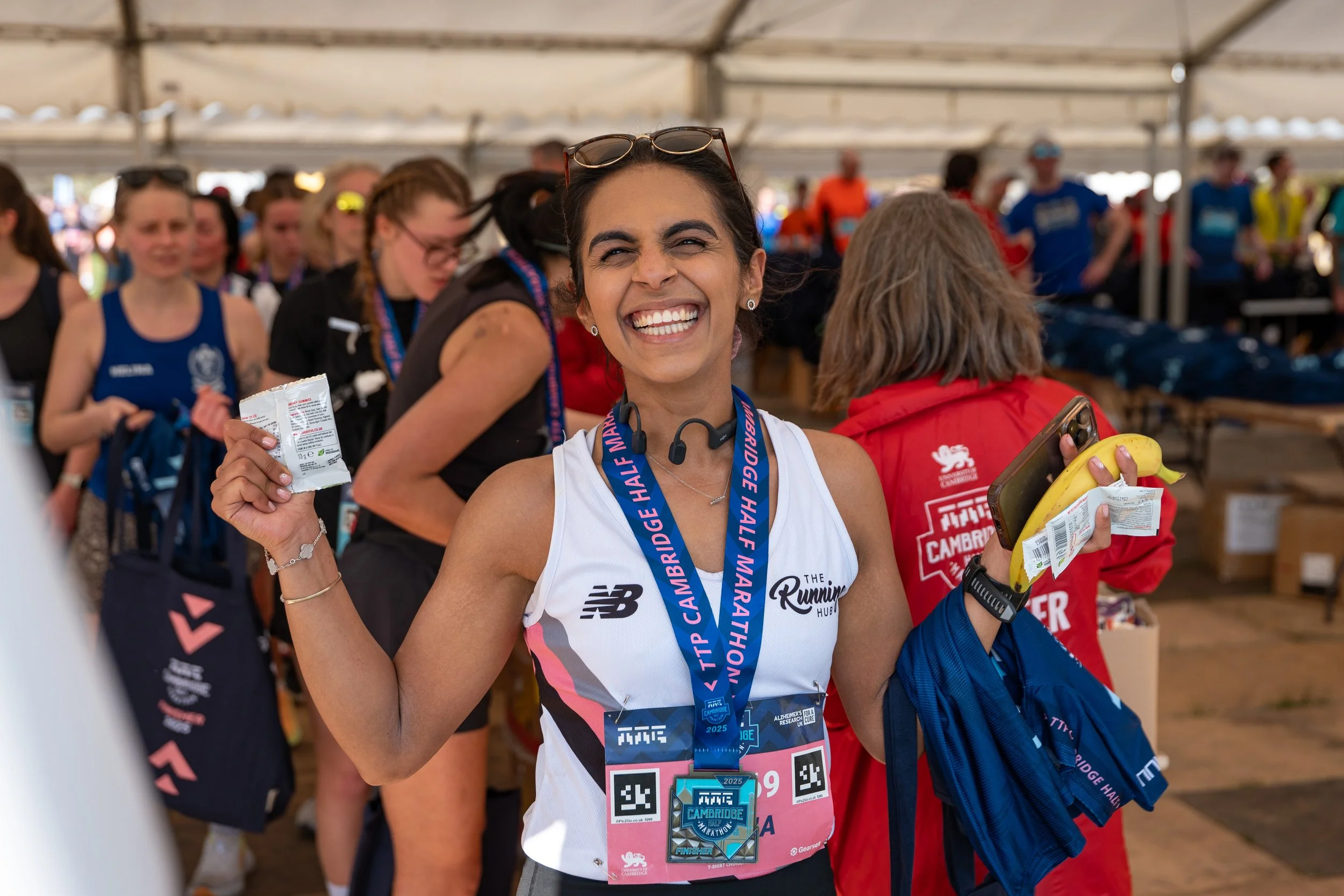 A woman smiling proudly at a marathon event, wearing a white sports top, a medal, and holding a banana snack, with other marathon participants in the background.