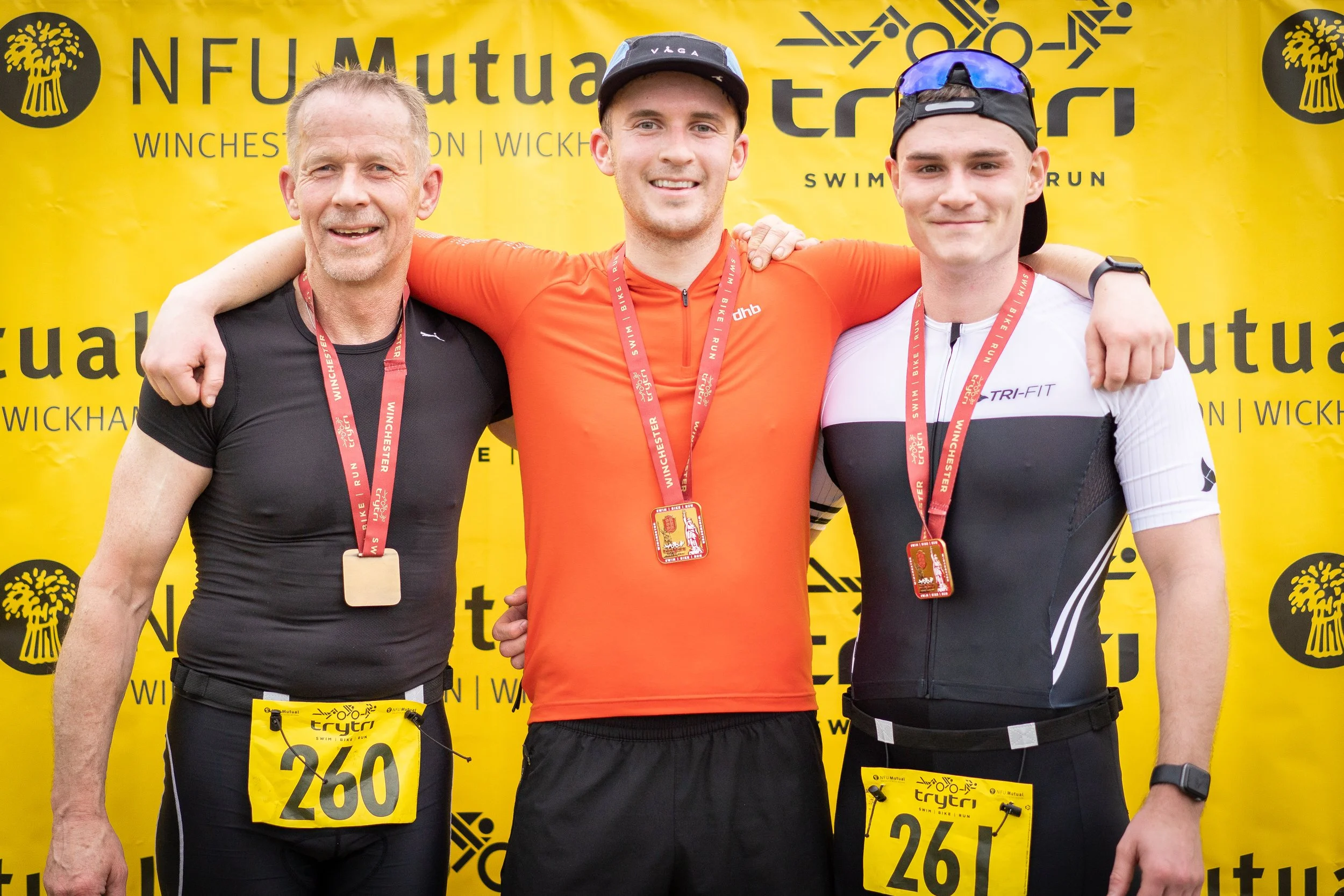 Three male marathon runners standing together with medals around their necks and race bibs, smiling after finishing a race, with a yellow backdrop displaying race event logos and sponsors.