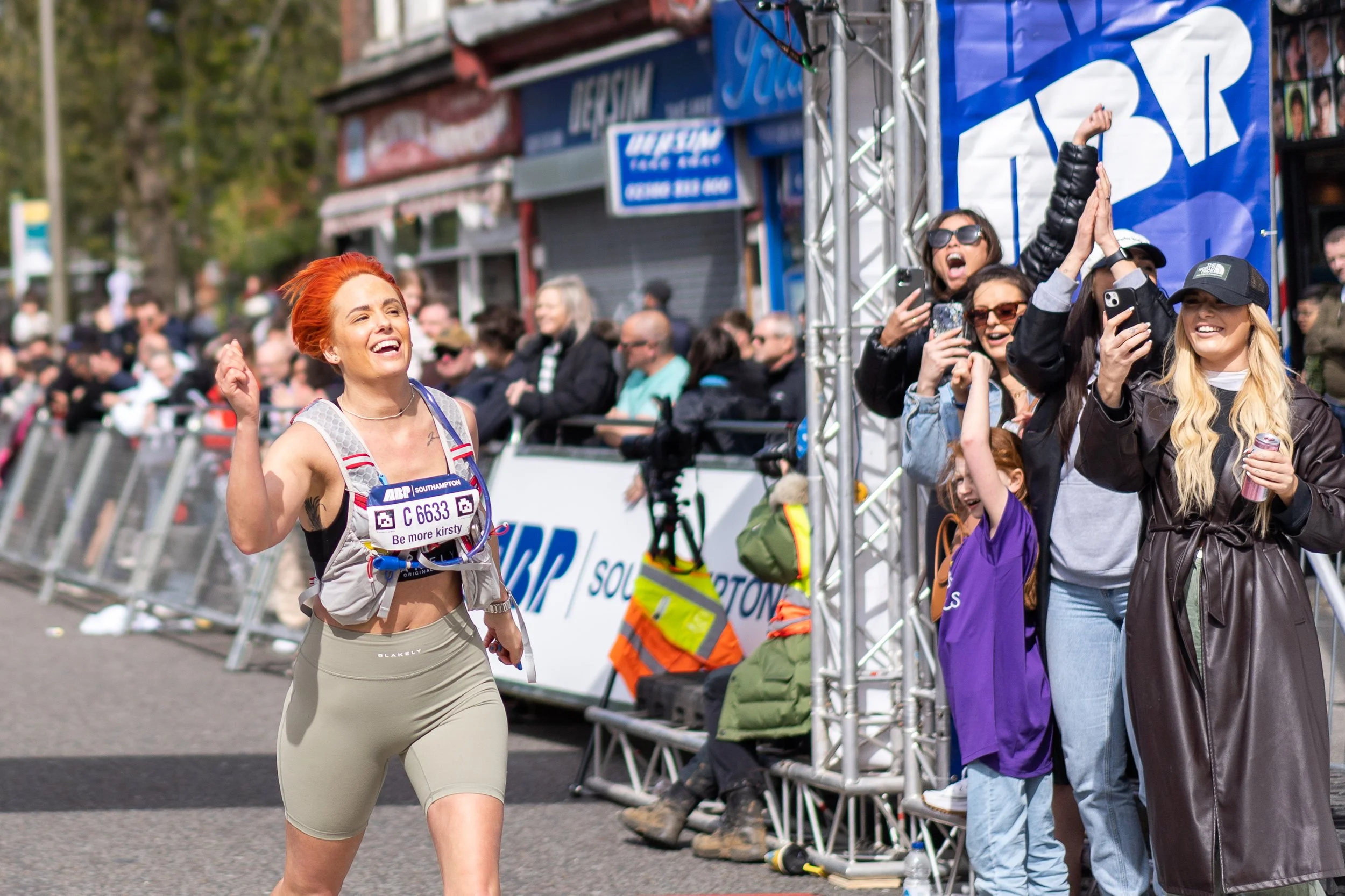 Female marathon runner smiling and celebrating as she crosses the finish line while spectators cheer and take photos nearby.