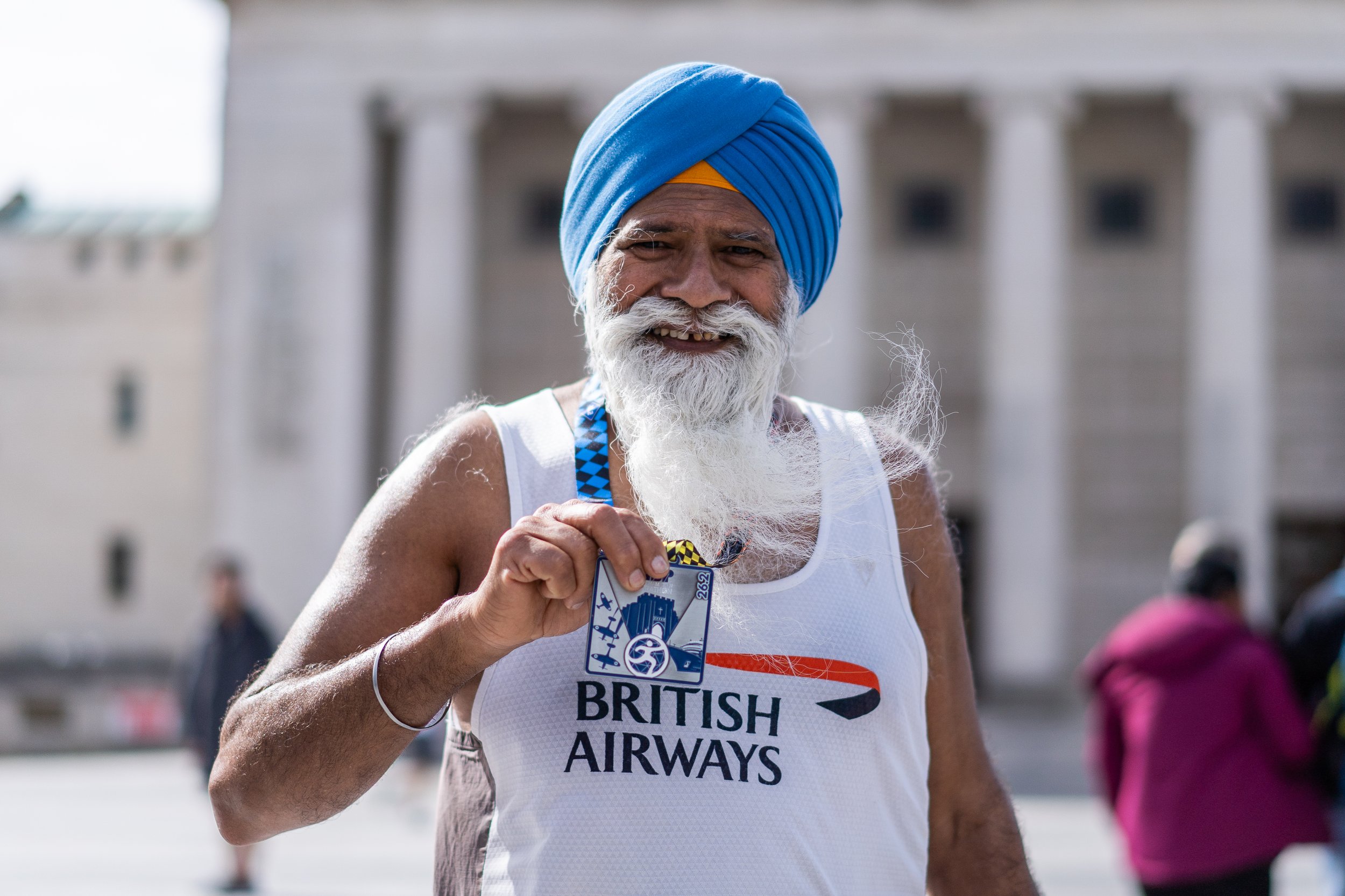 An elderly man with a white beard wearing a blue turban and a white athletic tank top with 'British Airways' logo, smiling and holding a finisher medal, standing outdoors near a government building.