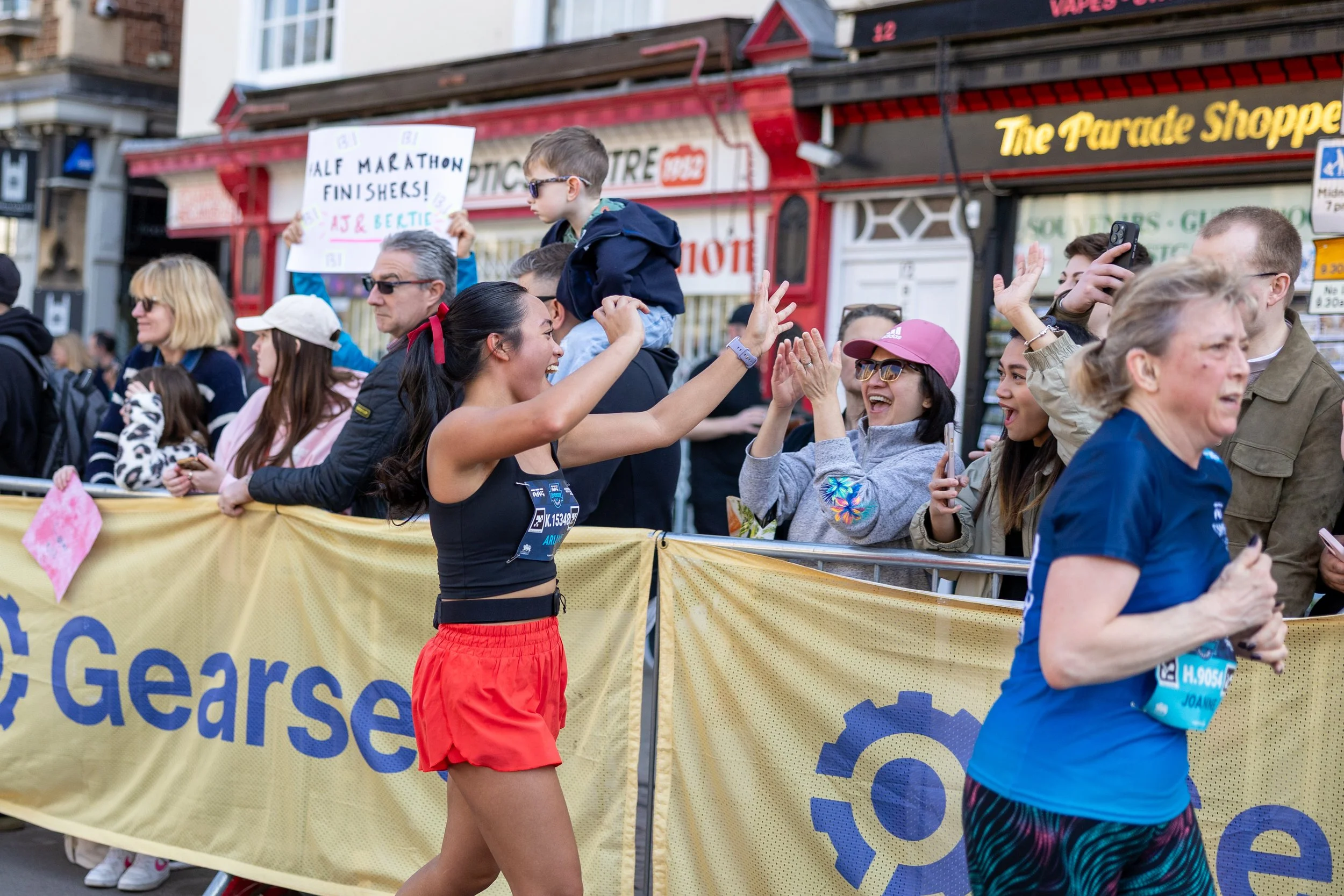 A woman runner in red shorts high-fives spectators at the finish line of a marathon while other runners and spectators are visible in the background.