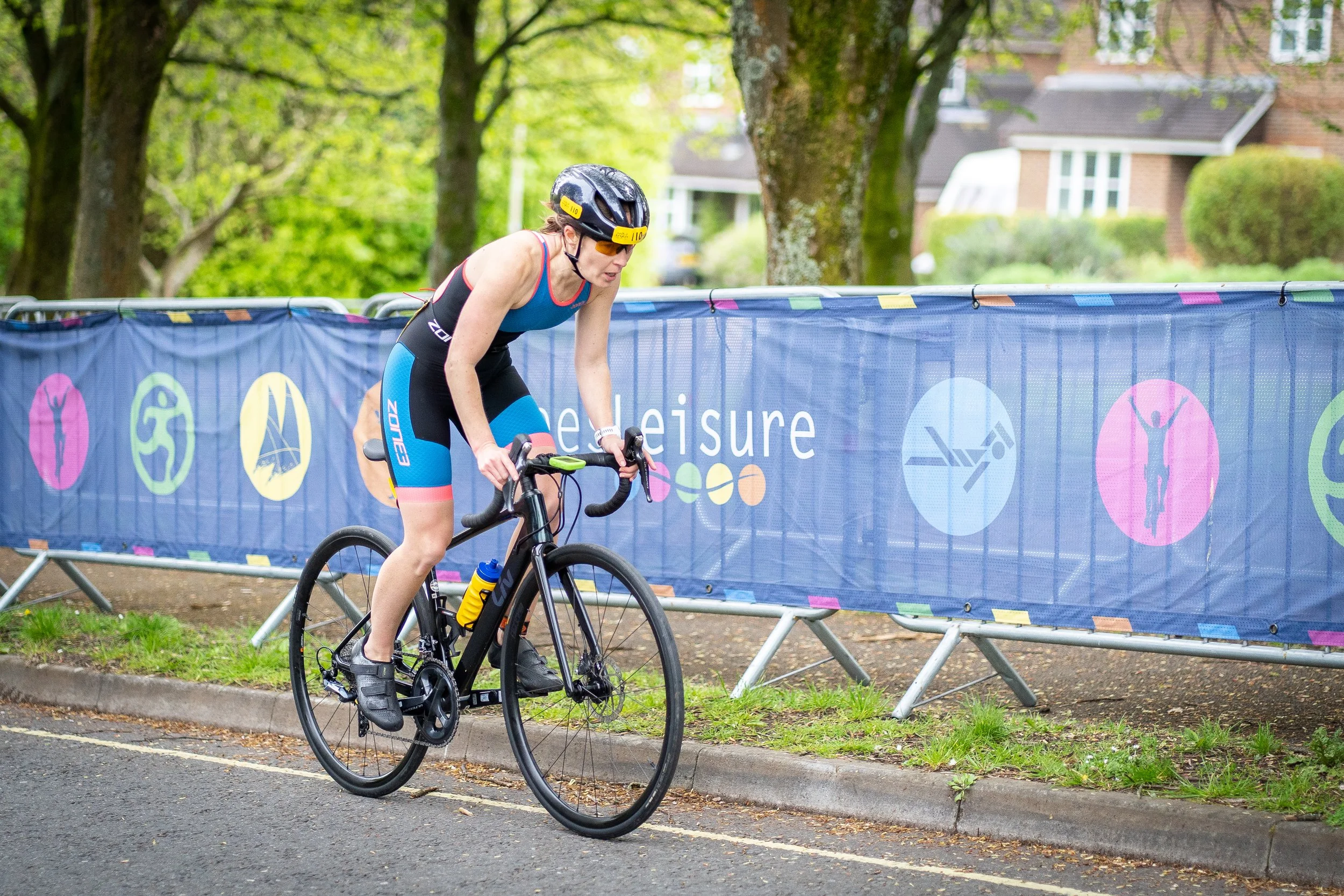 A woman in a black and blue triathlon suit riding a black road bike with a yellow water bottle, wearing a black helmet, on a street lined with trees and a colorful safety barrier.
