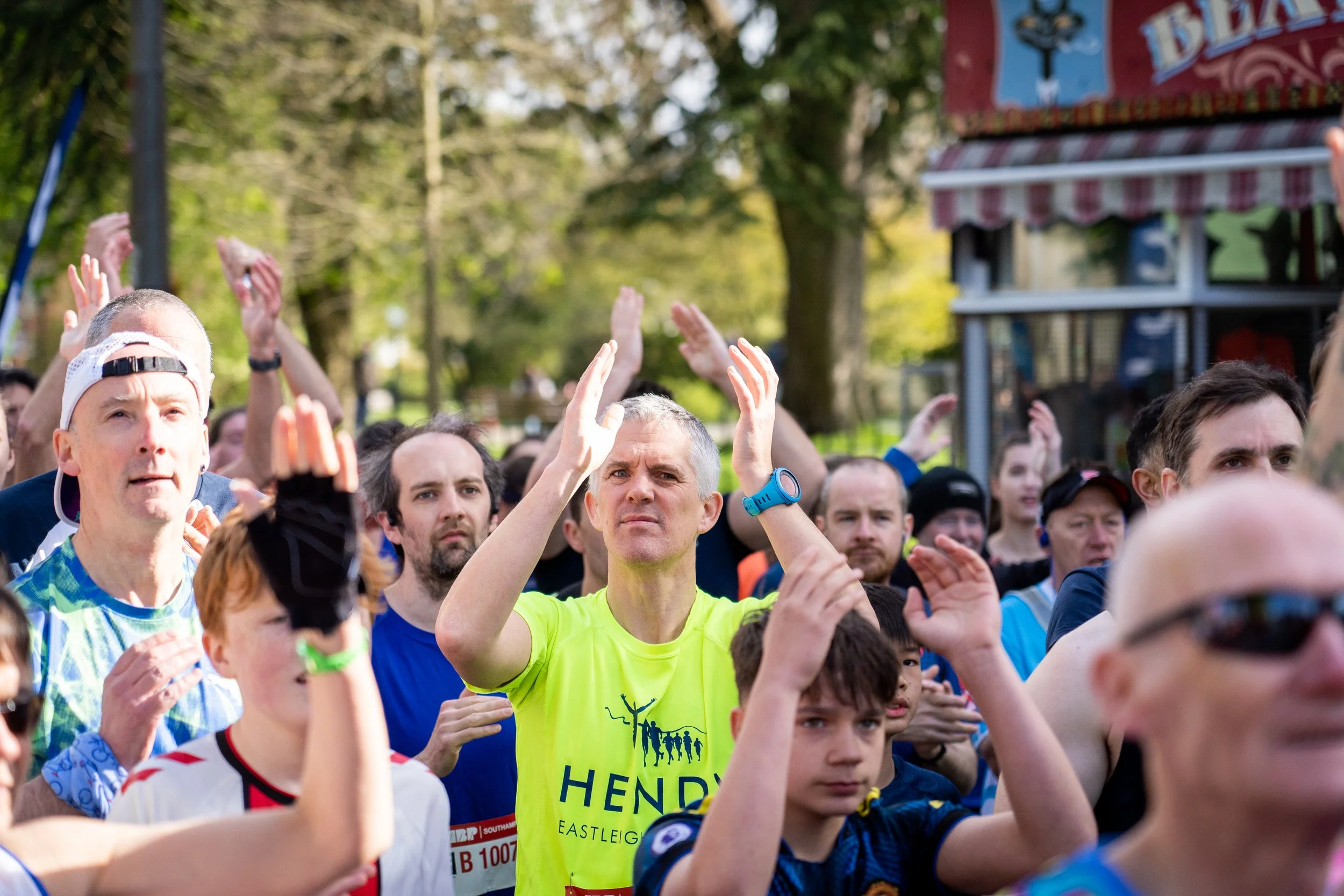 A crowd of people participating in a running event outdoors, raising their hands and clapping, with trees and a red and white striped tent in the background.