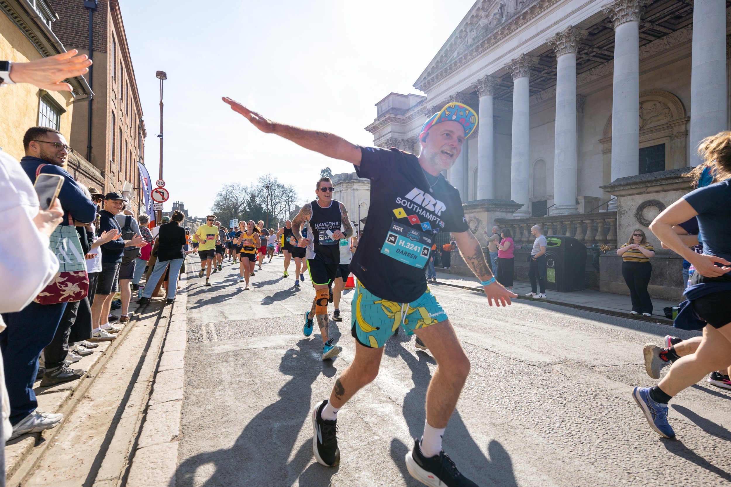 A man with a colorful hat and shorts running in a marathon, making a playful gesture with arms extended, surrounded by spectators and other runners on a city street with a historic building in the background.