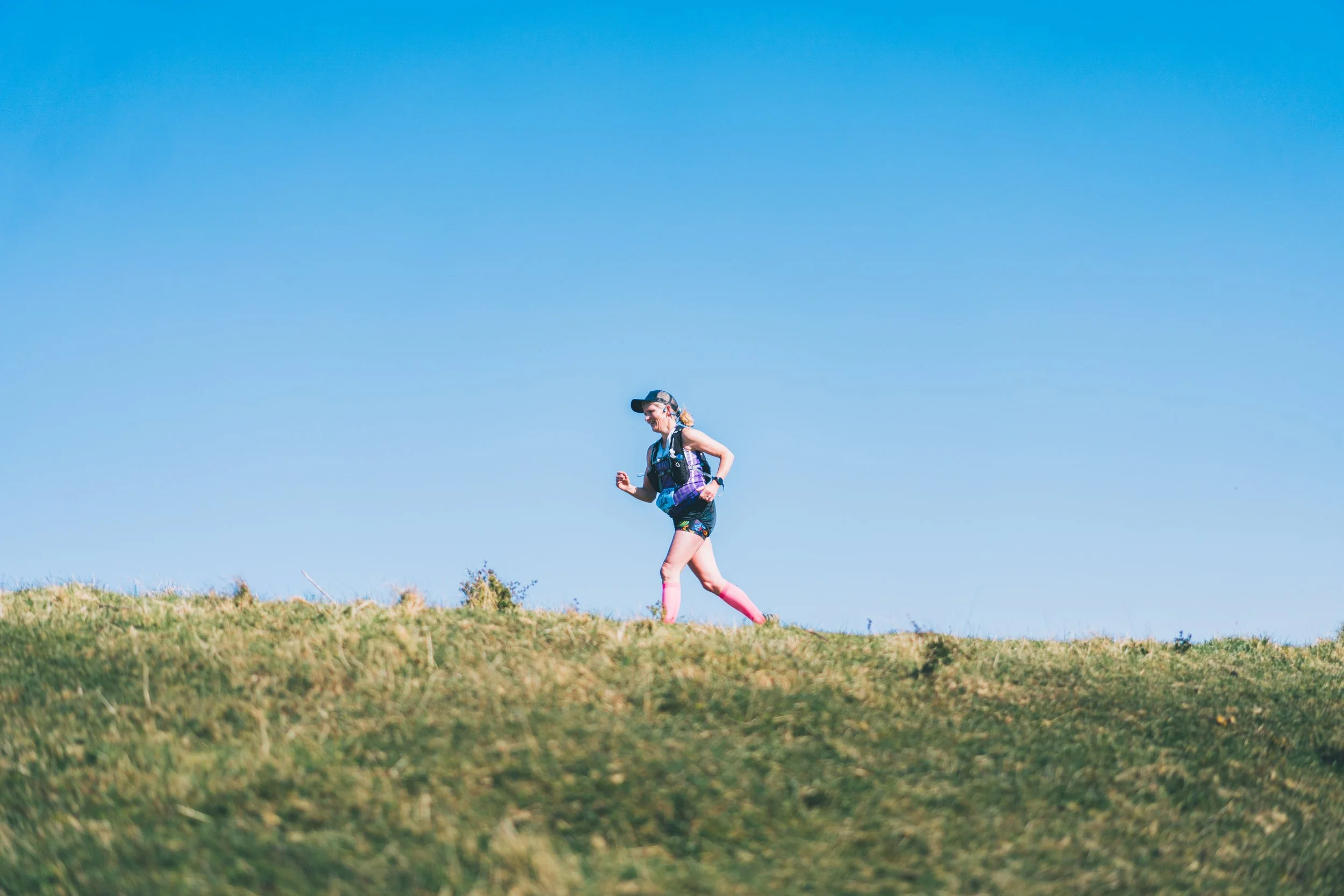Woman running outdoors on grassy hill, wearing sports attire, pink socks, cap, and sunglasses.