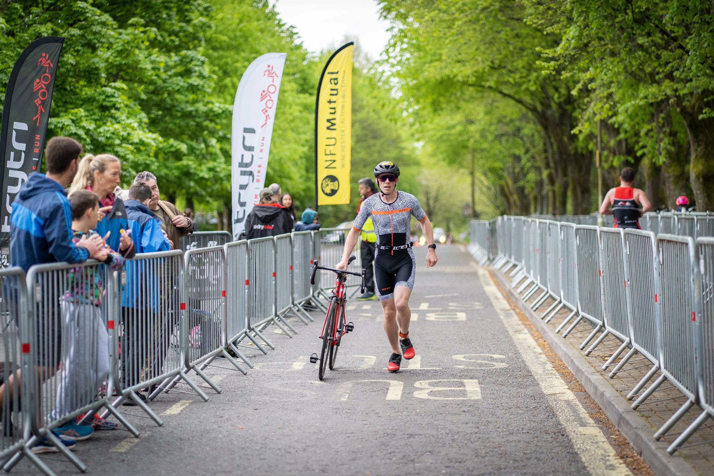 Triathlete running while holding a bike in a park during a race, with spectators on the side behind barriers, and trees lining the path.