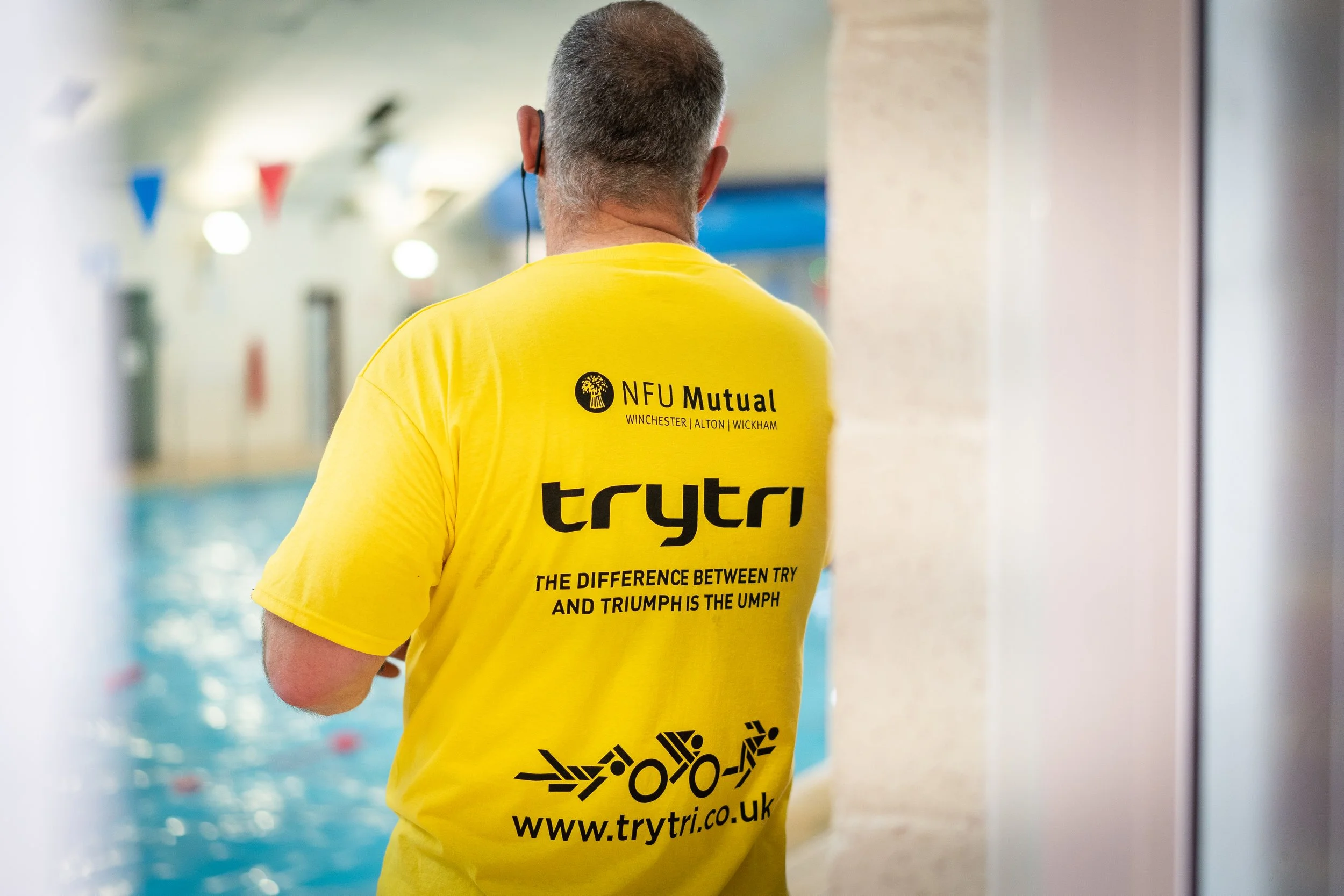 Back of a man wearing a yellow Trytri T-shirt, standing near an indoor swimming pool, with swim lanes and pool flags visible in the background.