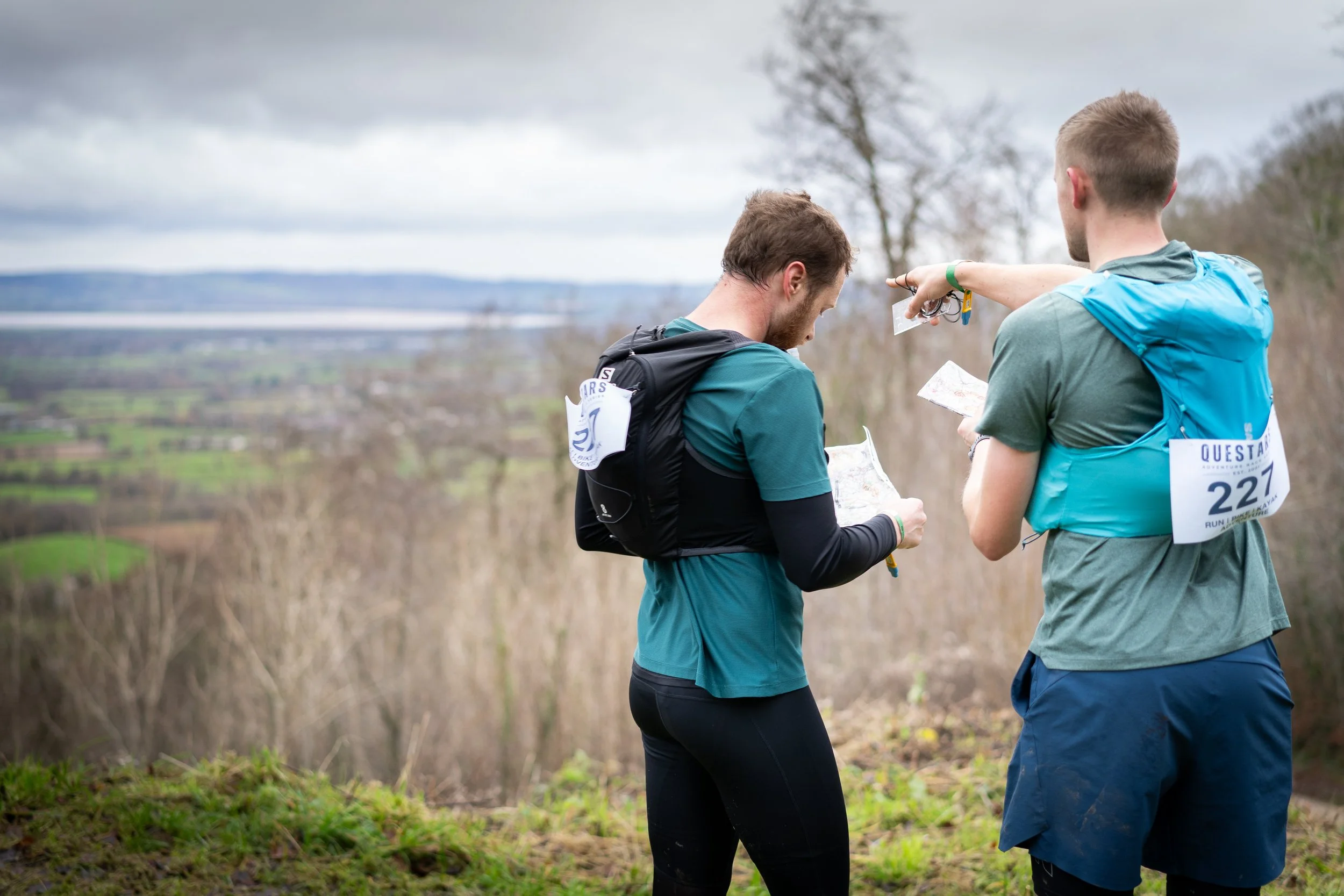 Two male trail runners in athletic gear checking maps outdoors on a cloudy day, with a scenic landscape of hills and fields in the background.