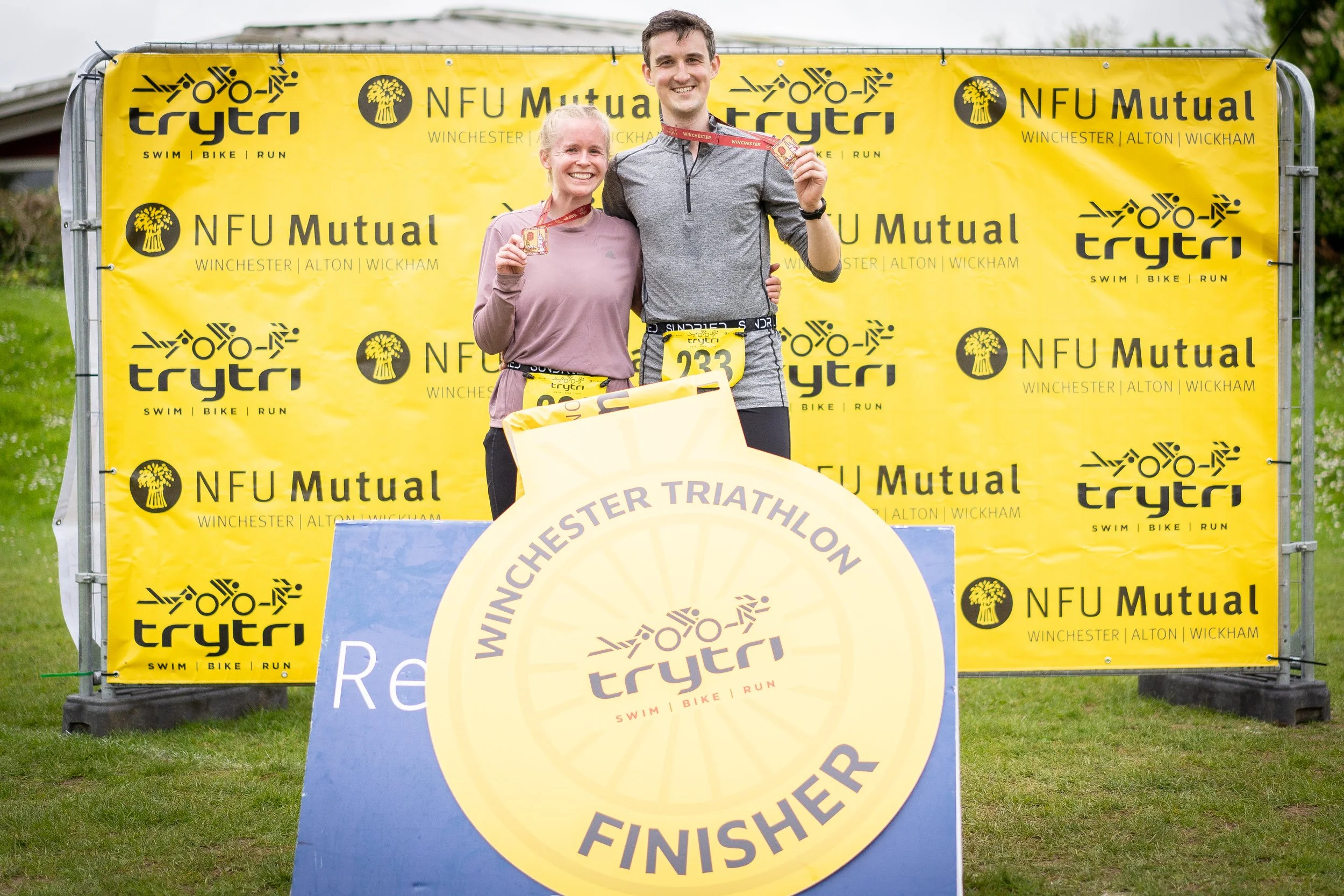 Two smiling race finishers with medals, standing behind a large yellow circular sign that reads "Winchester Triathlon Finisher". The background features a yellow banner with black logos and text for "NFU Mutual" and "trytri" services.