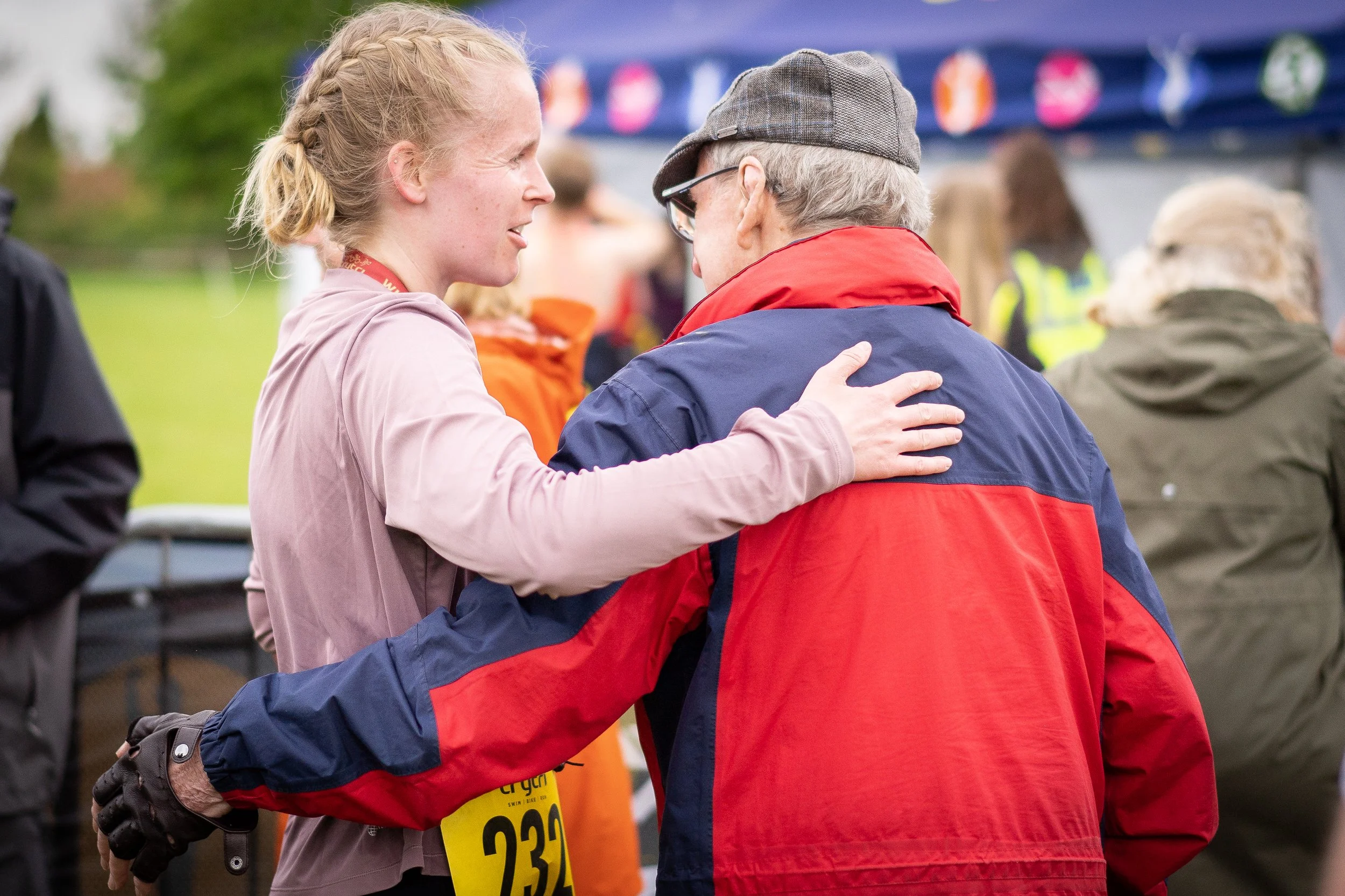 A young woman with a blonde braid talking to an older man wearing a gray cap and sunglasses at an outdoor event. The woman has her arm around the man's shoulder. Both are dressed in athletic or casual outdoor clothing, with the man in a red and blue